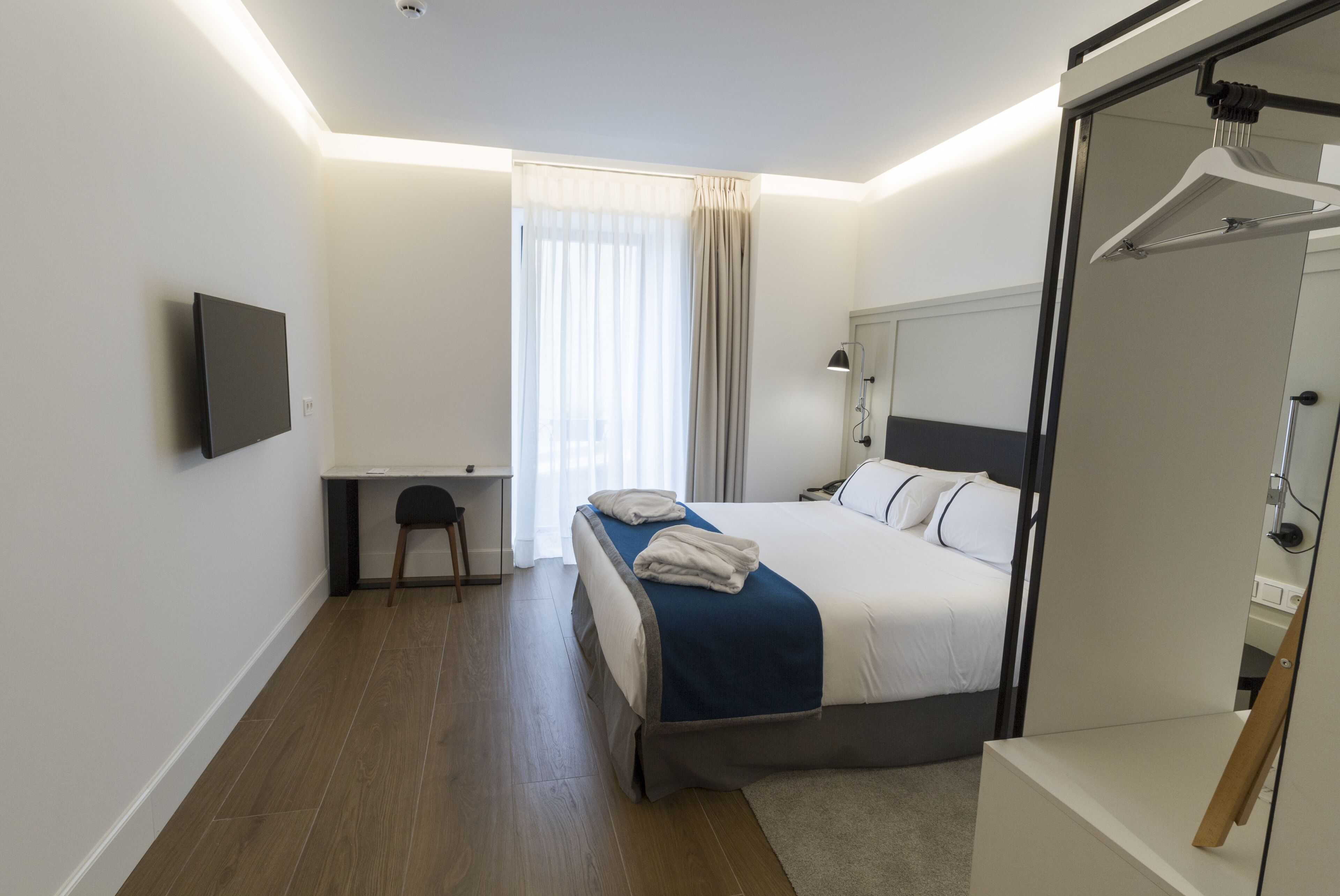 A modern hotel room with a bed, desk, chair, and a TV mounted on the wall.