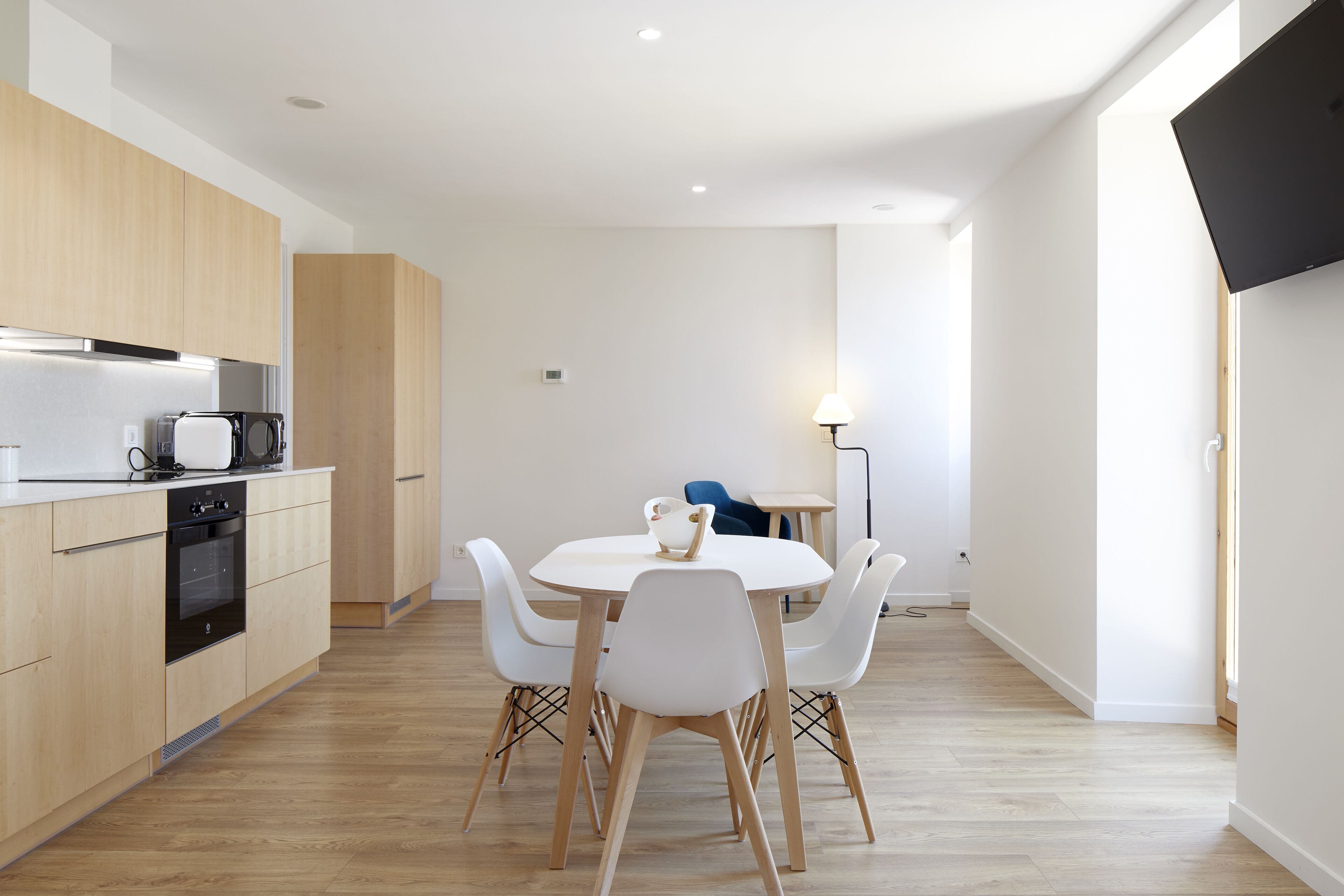 A modern kitchen with wooden cabinets, a built-in oven, and a dining area with a round table and white chairs.