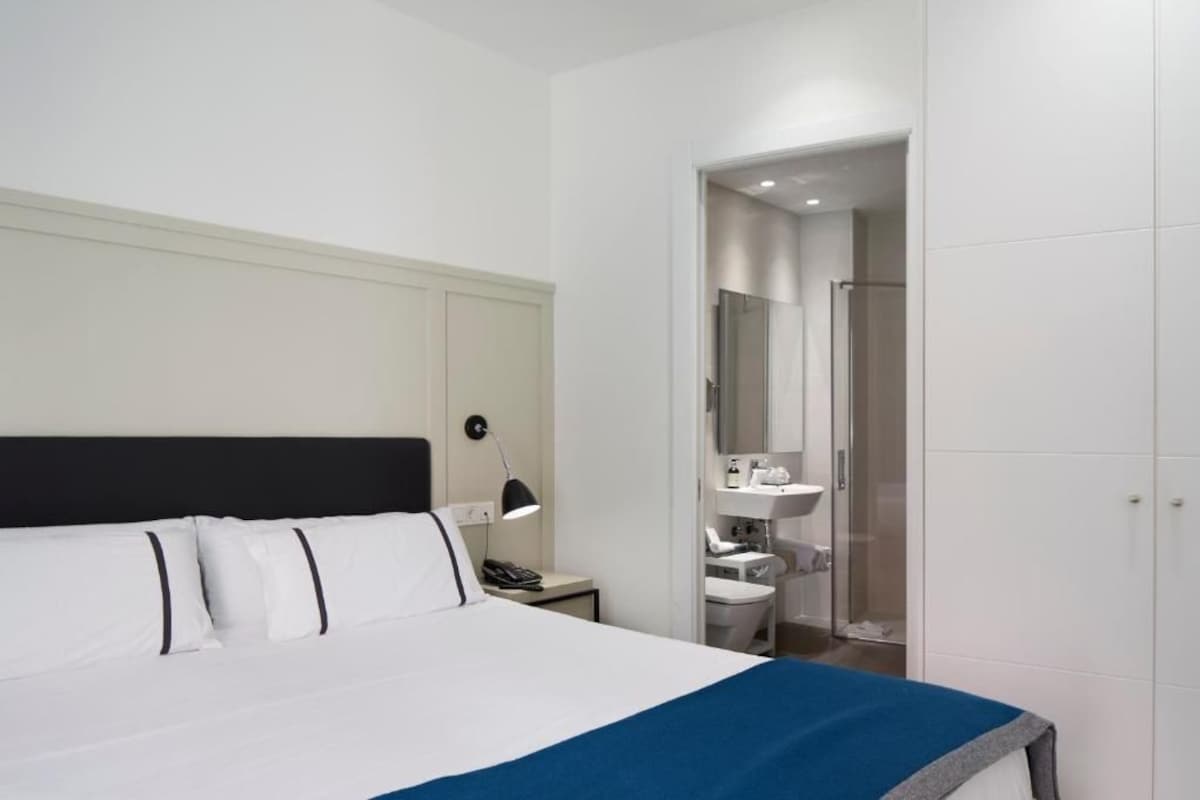 A hotel room with a bed, bedside table, lamp, and a bathroom with a sink and mirror.