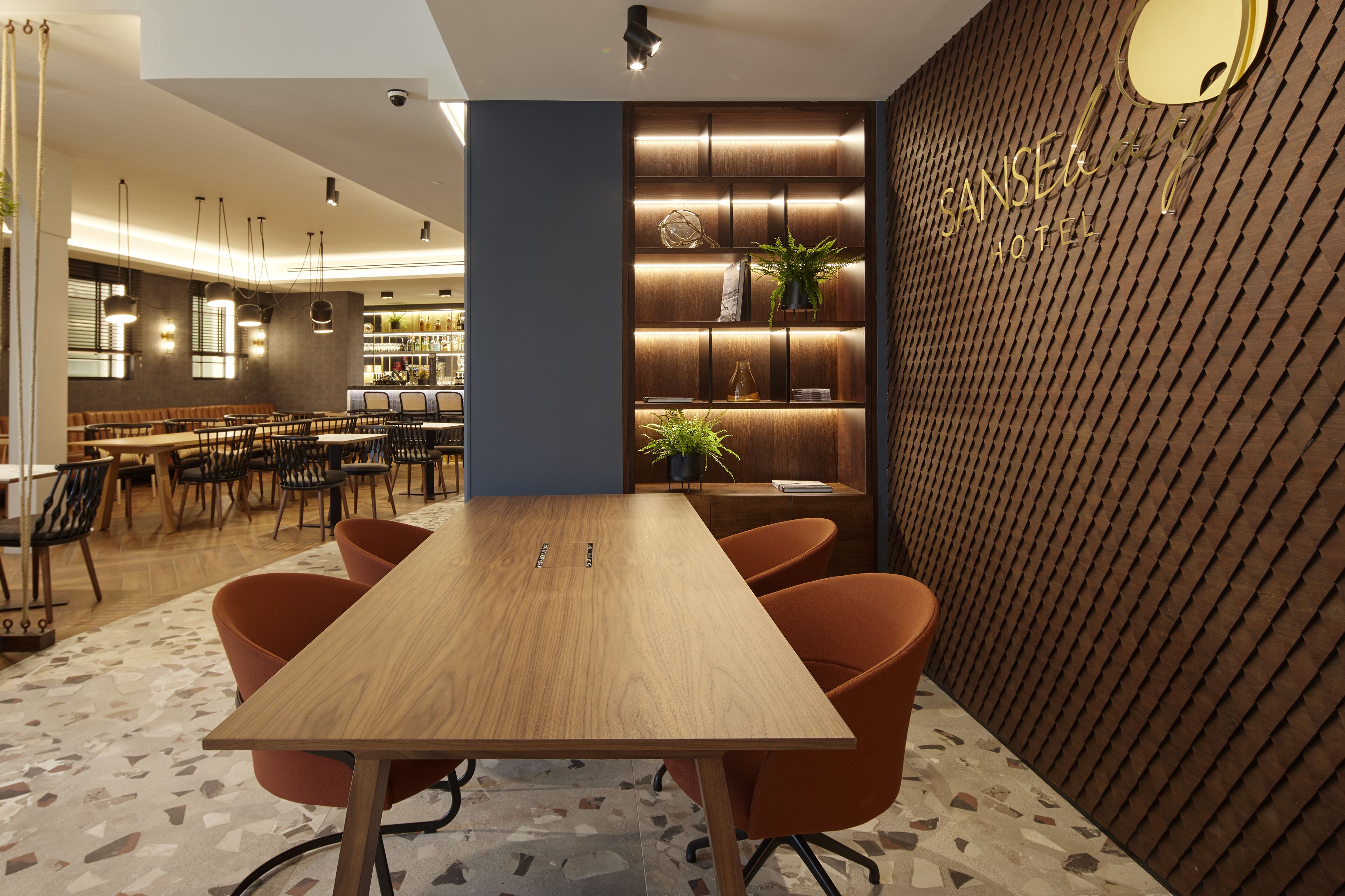 A modern hotel lobby with a wooden reception desk, orange chairs, a bar area, and a patterned wall with the hotel name.