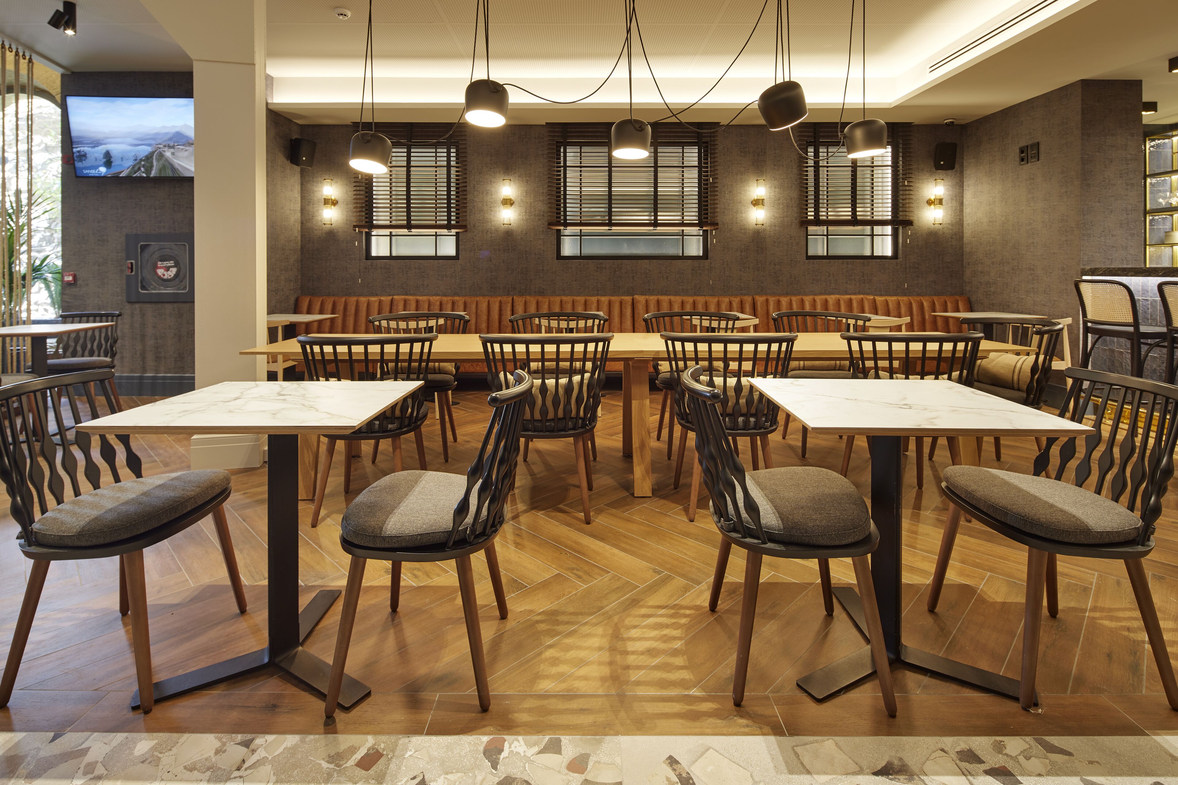 A restaurant with multiple tables and chairs, a television mounted on the wall, and a herringbone-patterned floor.