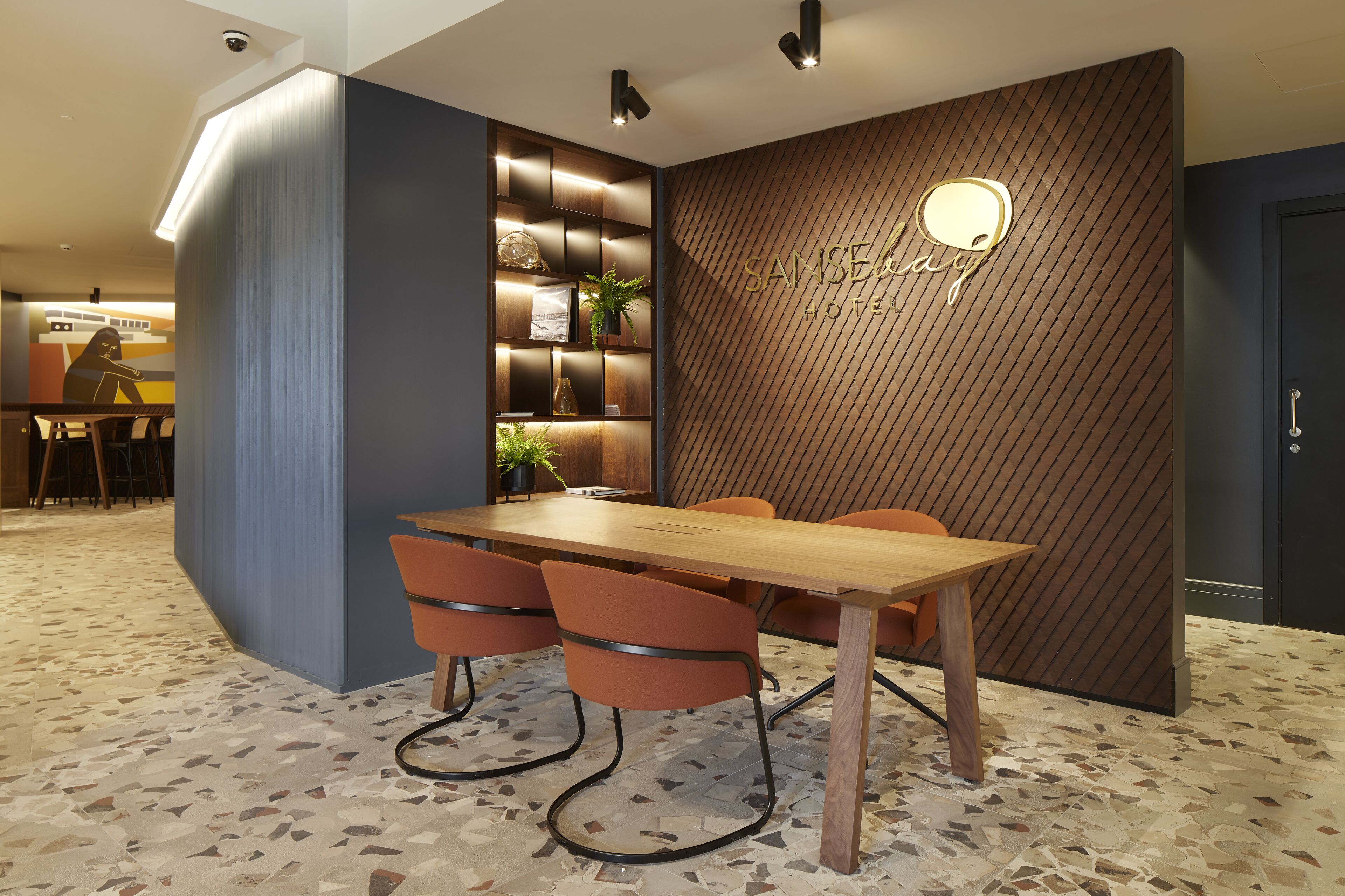A modern hotel lobby with a wooden reception desk, a seating area with orange chairs, and a patterned floor.
