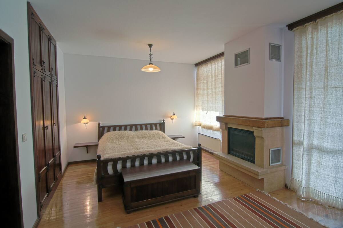 A bedroom with a bed, bench, fireplace, and wall-mounted lights.