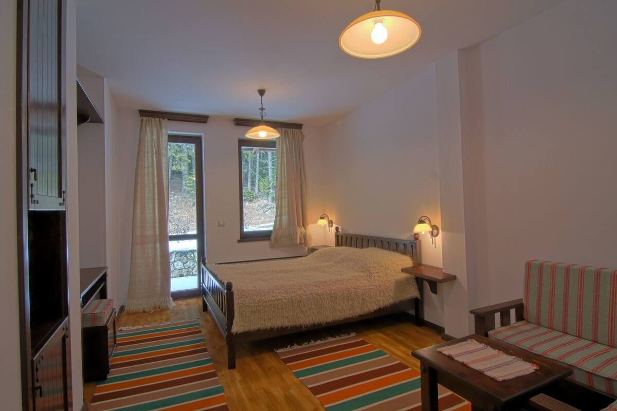 A bedroom with a bed, a sofa, a wooden floor, a window with curtains, and a hanging light fixture.