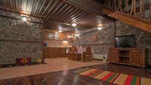 A rustic interior with a stone wall, wooden ceiling beams, a dining area with a table and chairs, and a television on a wooden cabinet. - Holiday Village Kochorite (Smolyan)