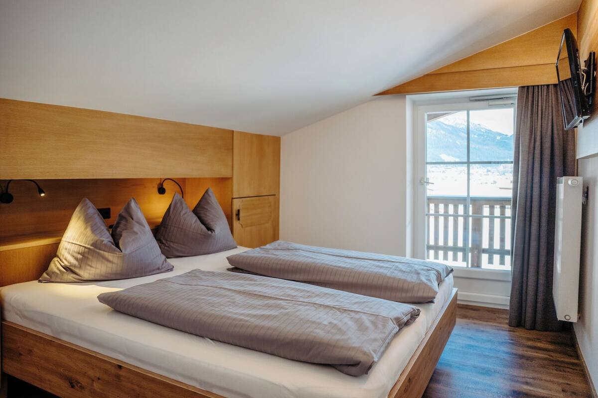 A bedroom with two beds, wooden headboard, a window with a view of mountains, and a balcony with a railing.