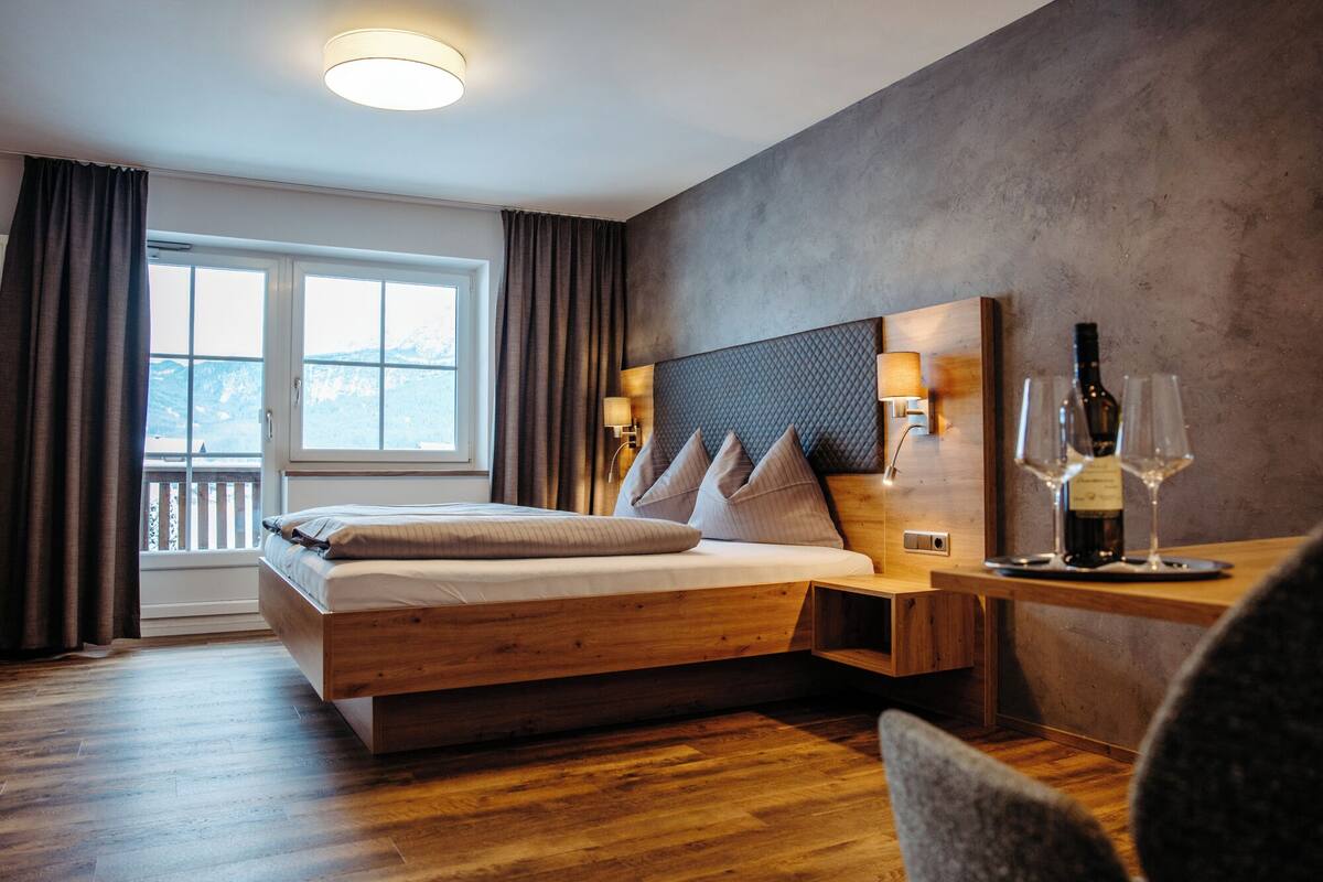A modern hotel room with a wooden bed, a nightstand with a wine bottle and glasses, and a view of mountains through the window.