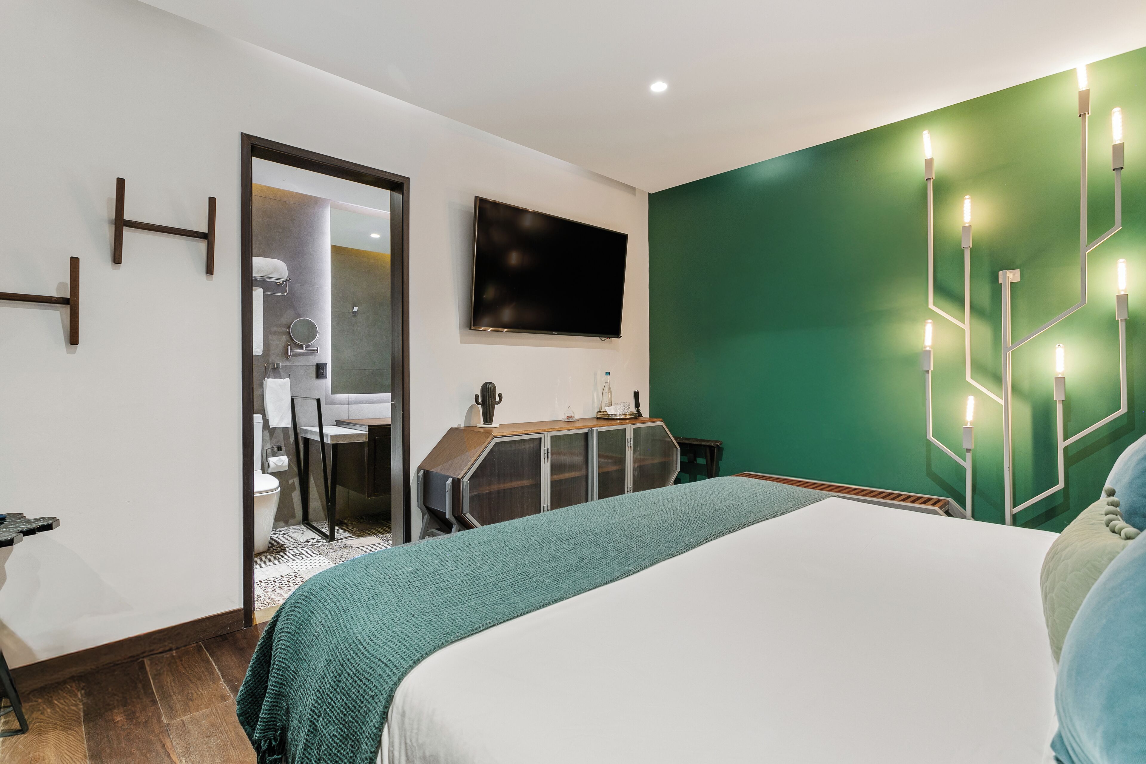 A modern hotel room with a bed, a desk, a mirror, a television, and a green accent wall.