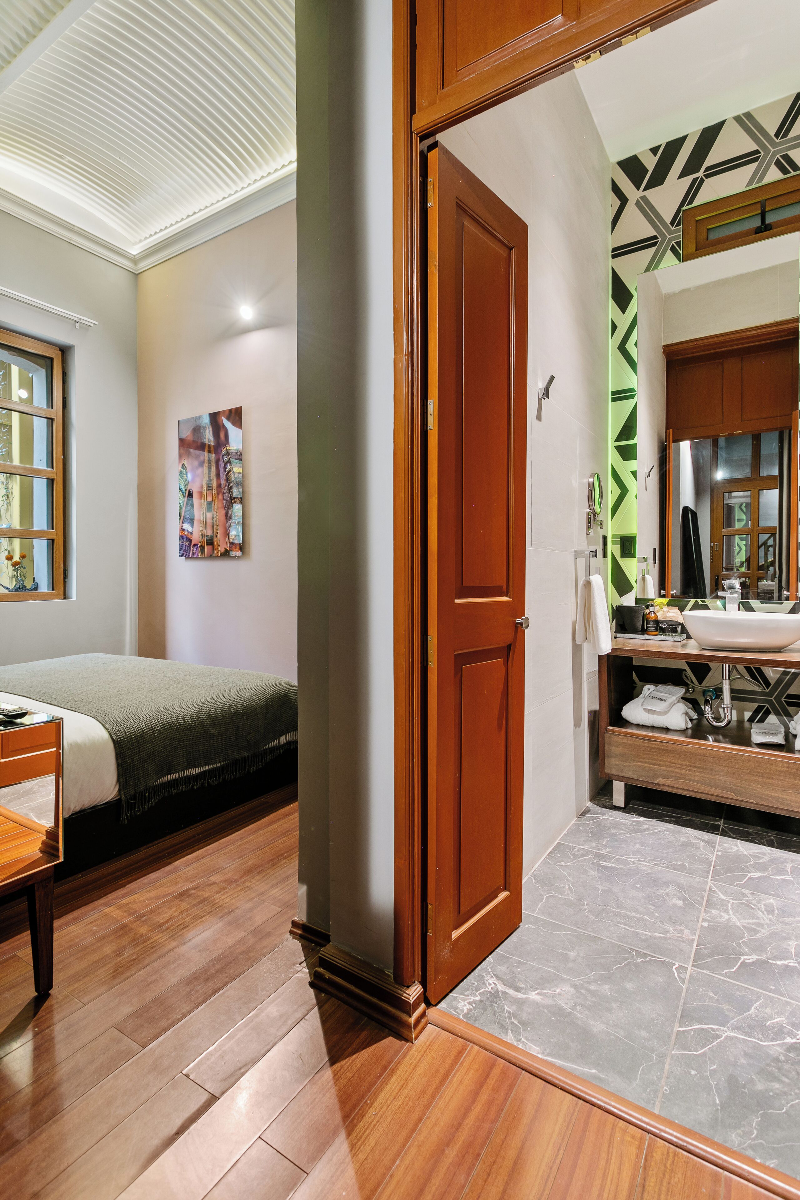 A bedroom with a wooden bed, a bedside table, a window, and a bathroom with a sink, mirror, and towel rack.