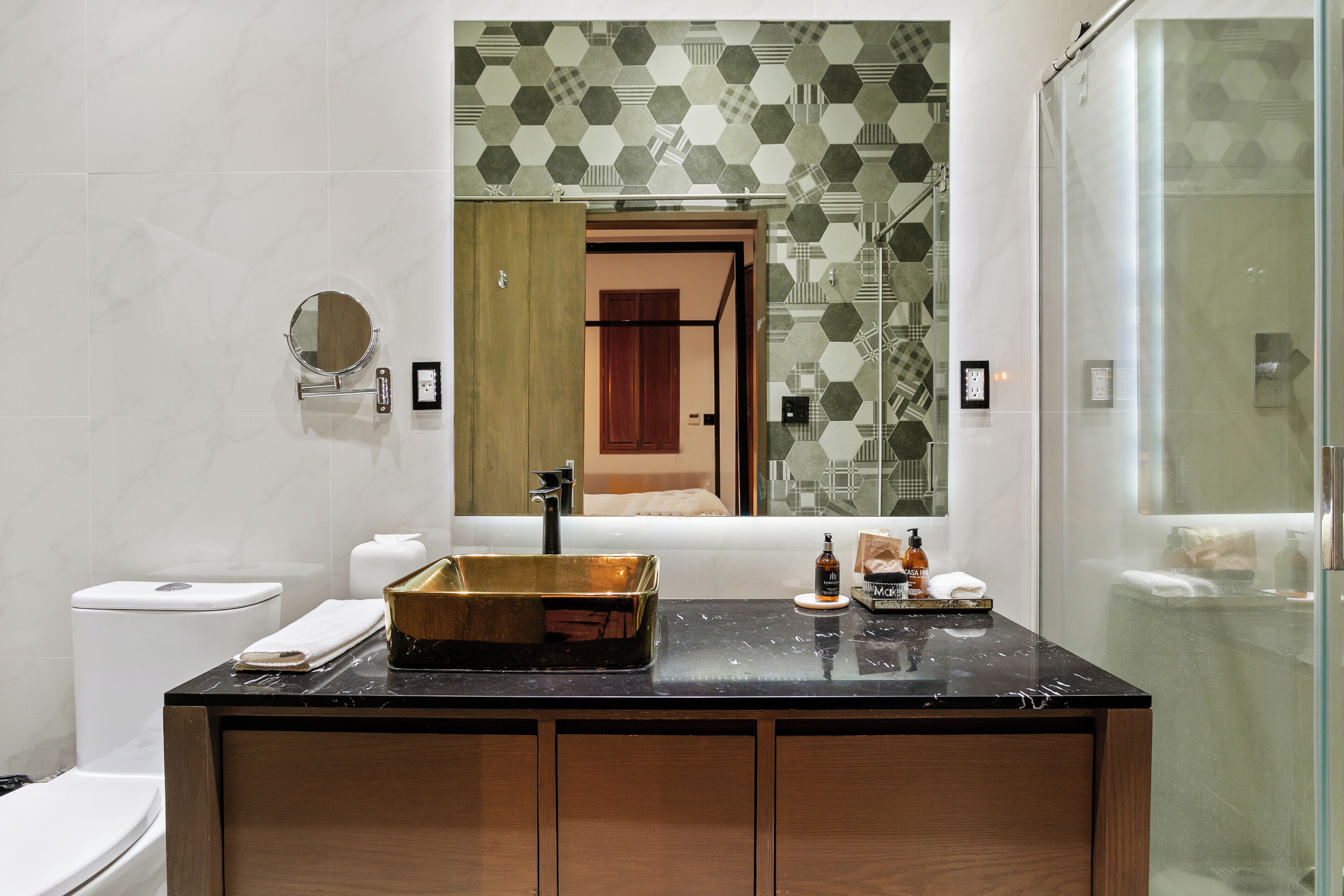 Un baño con encimera de mármol negro, lavabo dorado, espejo redondo y zona de ducha.