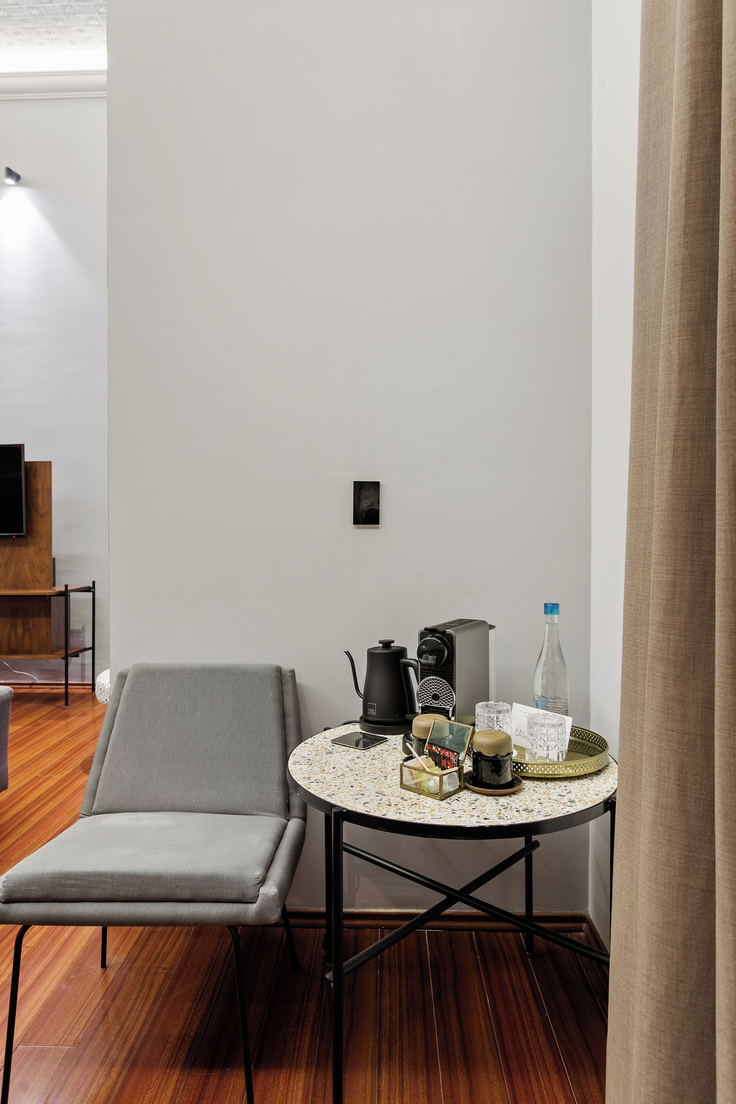 Una habitación pequeña y moderna con una silla gris, una mesa redonda con una cafetera, un hervidor y una botella de agua.