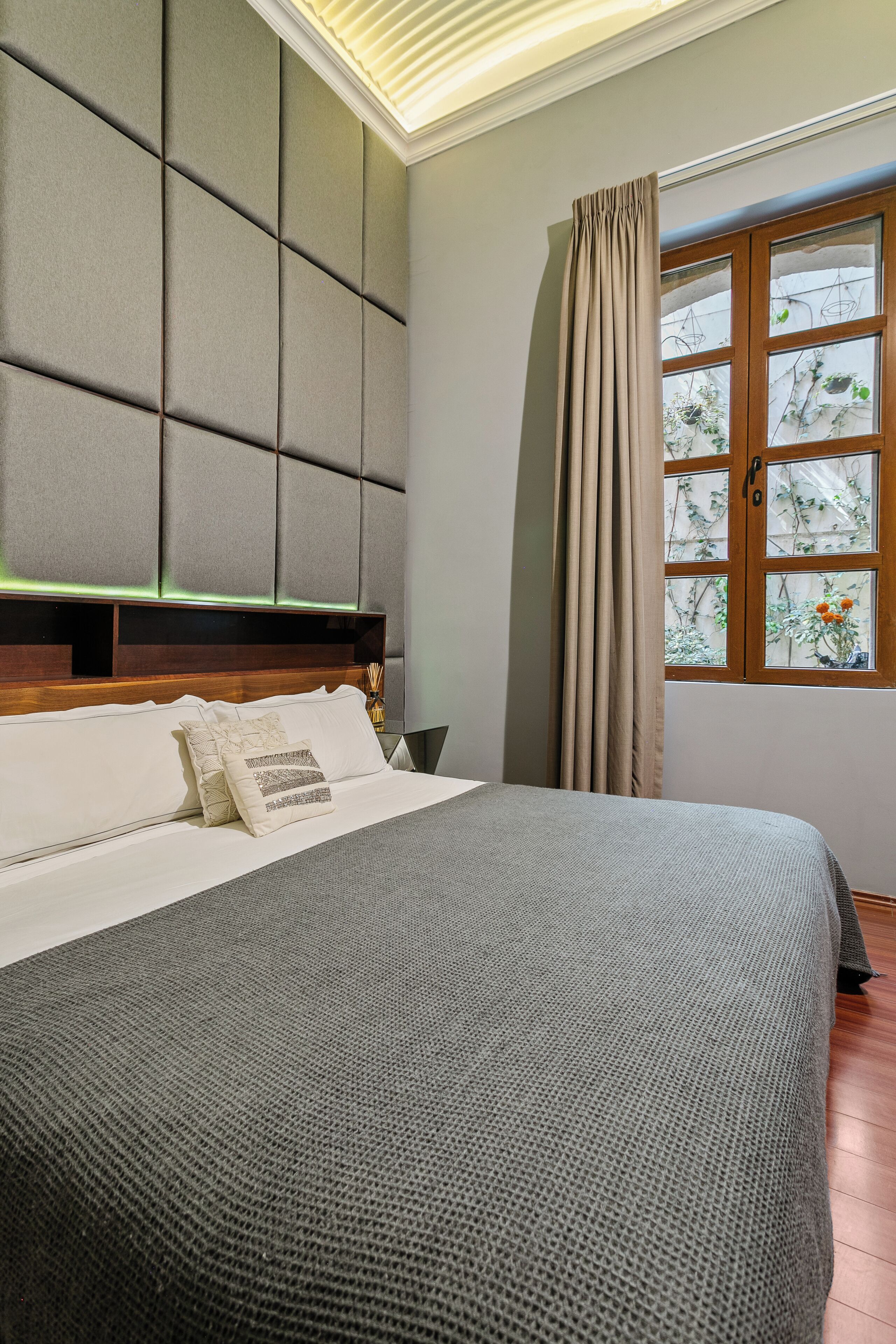 A bedroom with a bed, a headboard, a window with curtains, and a view of greenery outside.