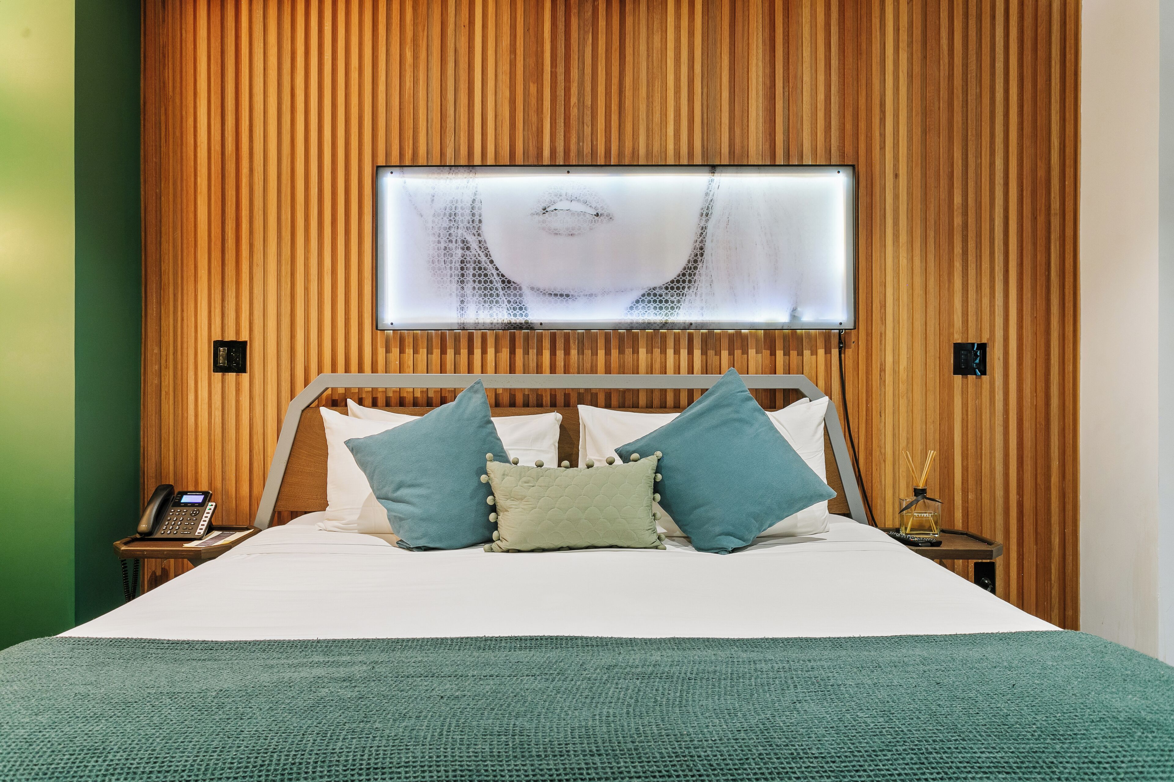 Un lit bien fait, avec une couverture verte, une tête de lit en bois et un téléviseur fixé au mur.