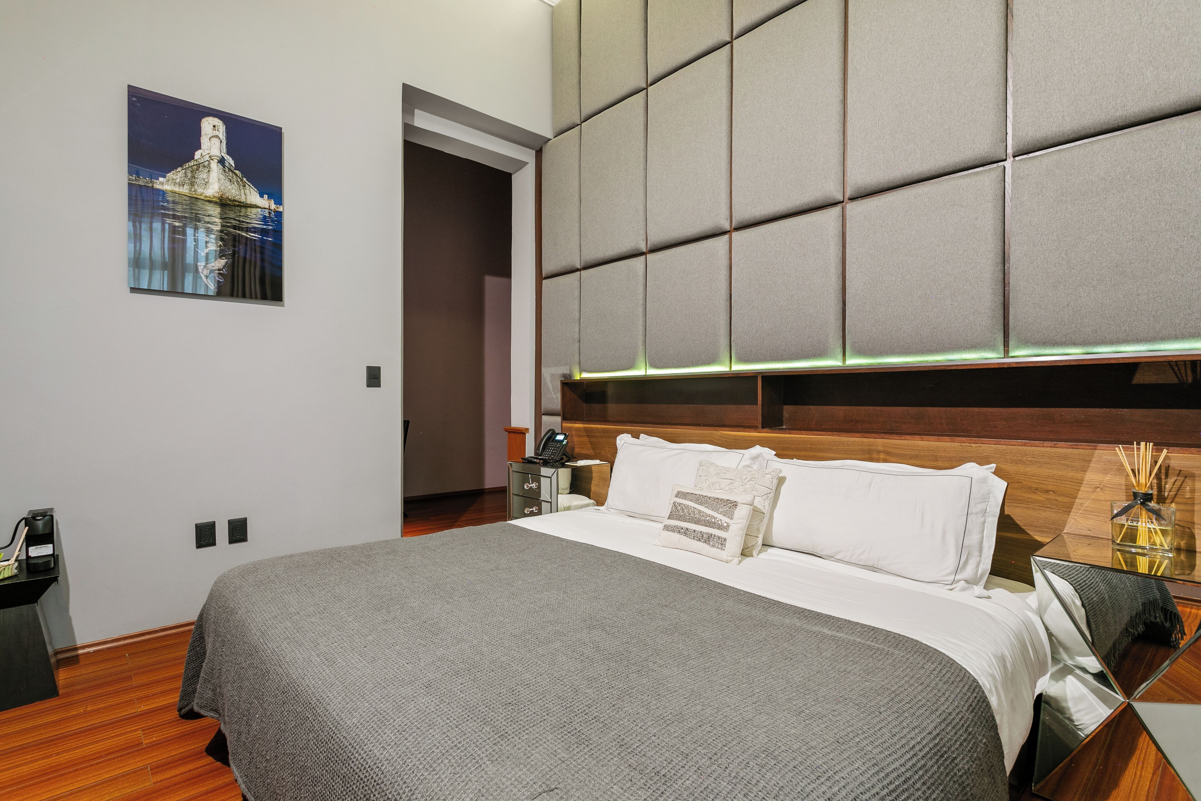 Una habitación de hotel moderna con una cama grande, un cabecero con diseño geométrico, una mesita de noche con lámpara y un cuadro en la pared.