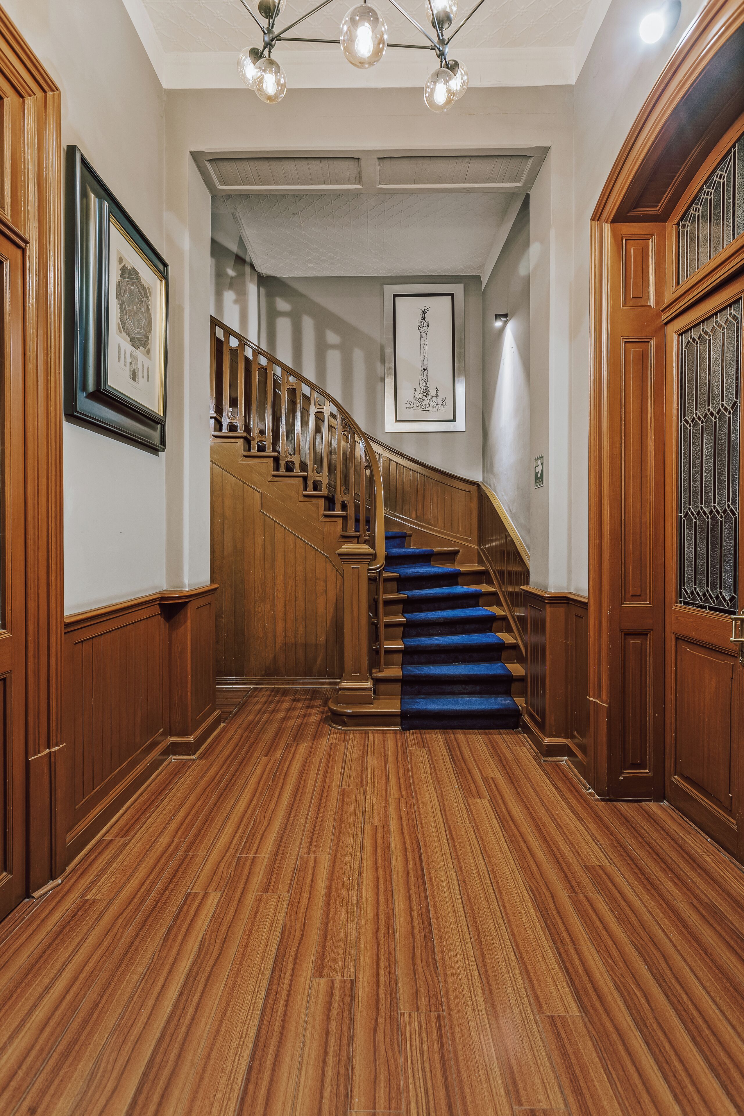 Una escalera de madera con alfombra azul que lleva a una plataforma, flanqueada por paneles de madera y una puerta con celosía metálica.