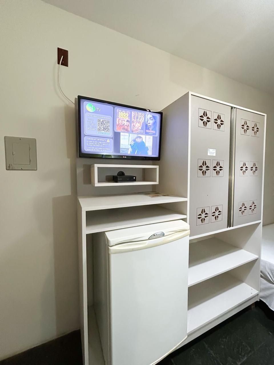 Ein Hotelzimmer mit einem Wand-Fernseher, einem weißen Schrank mit dekorativen Ausschnitten und einer weißen Minibar.