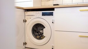 A compact, modern washing machine in a clean, white kitchen with a wooden countertop and a built-in cabinet. - Harbour House - Donnini Apartments (Ayr)