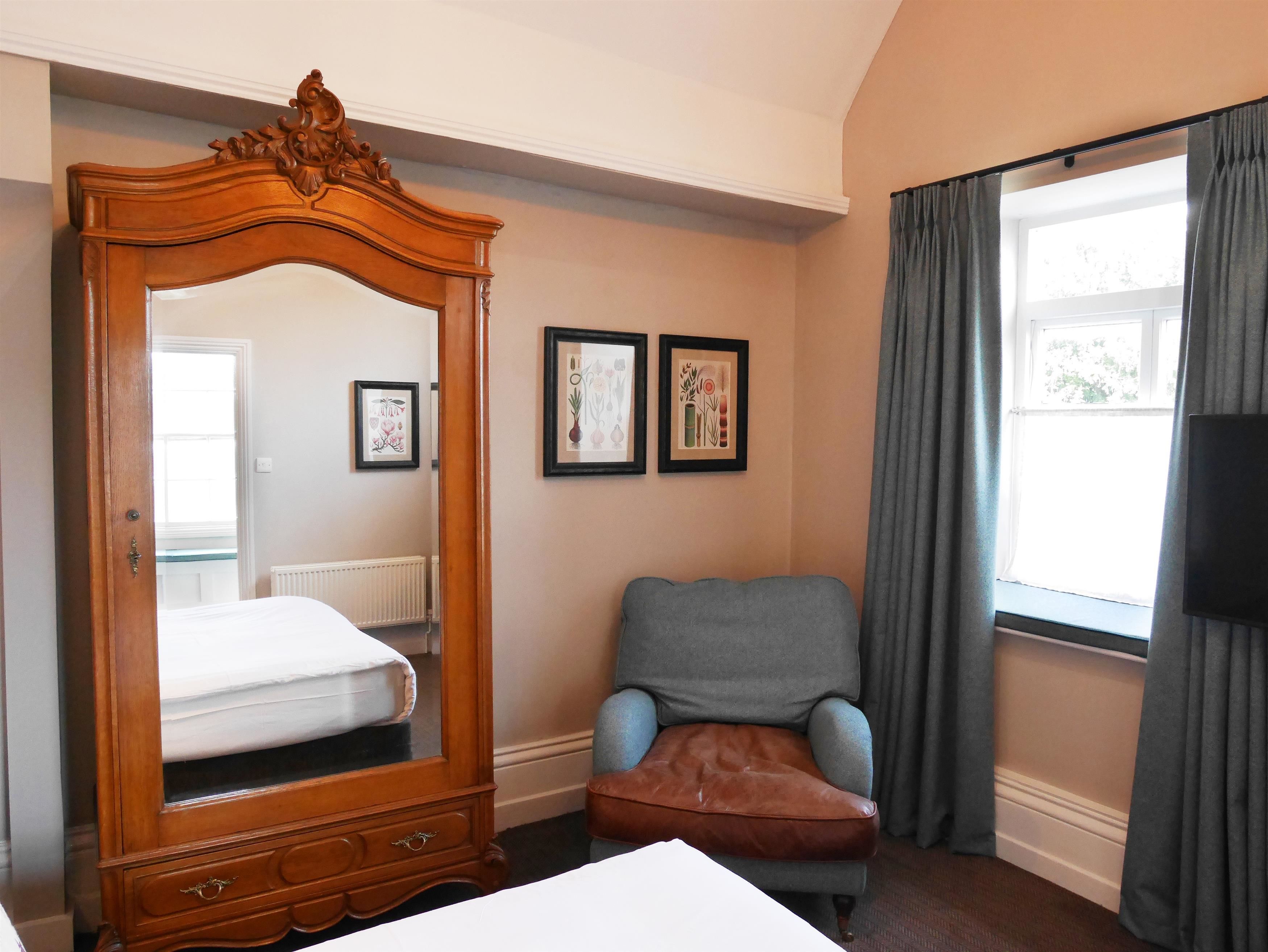 A bedroom with a wooden bed frame, a grey armchair, and a large mirror. There are two framed pictures on the wall and a window with curtains.
