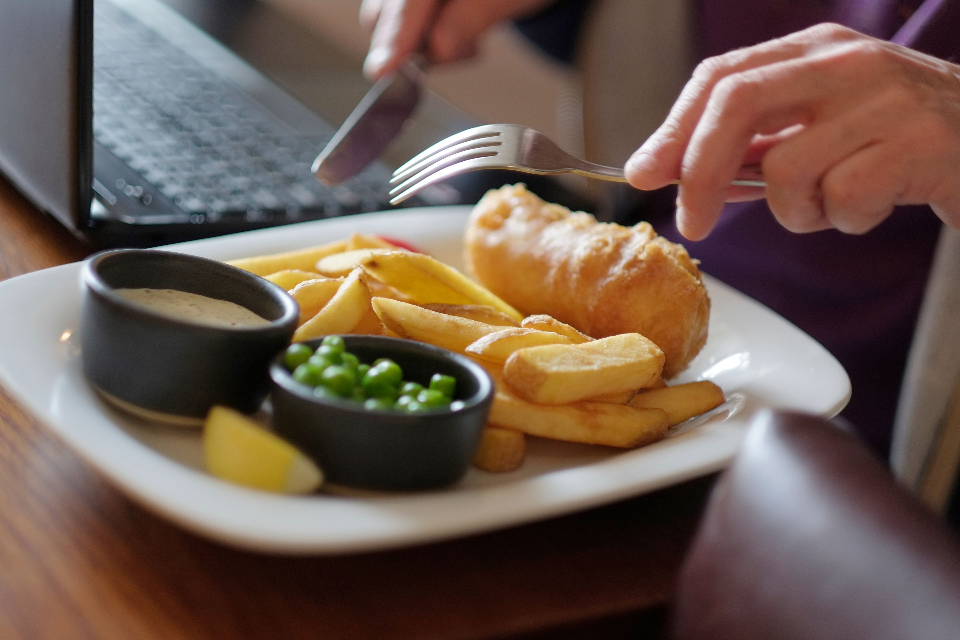 Un plato de comida con pescado, papas fritas, guisantes y salsa, y una computadora portátil al fondo.