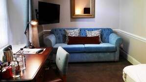 A hotel room with a blue sofa, a wooden desk, a chair, a TV, and a mirror on the wall. - The Fleece (Witney)