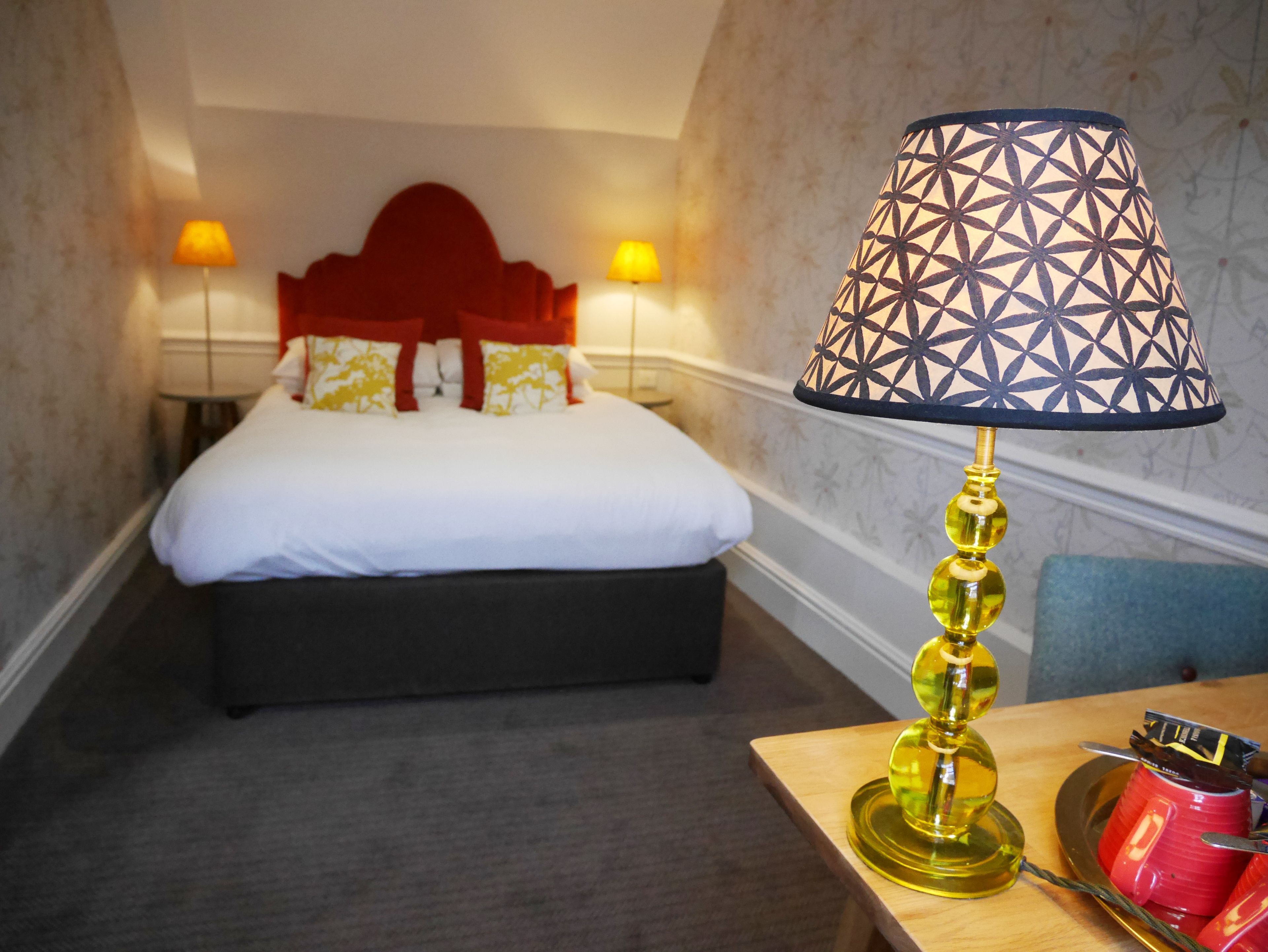 Une chambre d’hôtel avec un lit, deux lampes de chevet, une table de chevet avec une lampe et une tasse rouge.