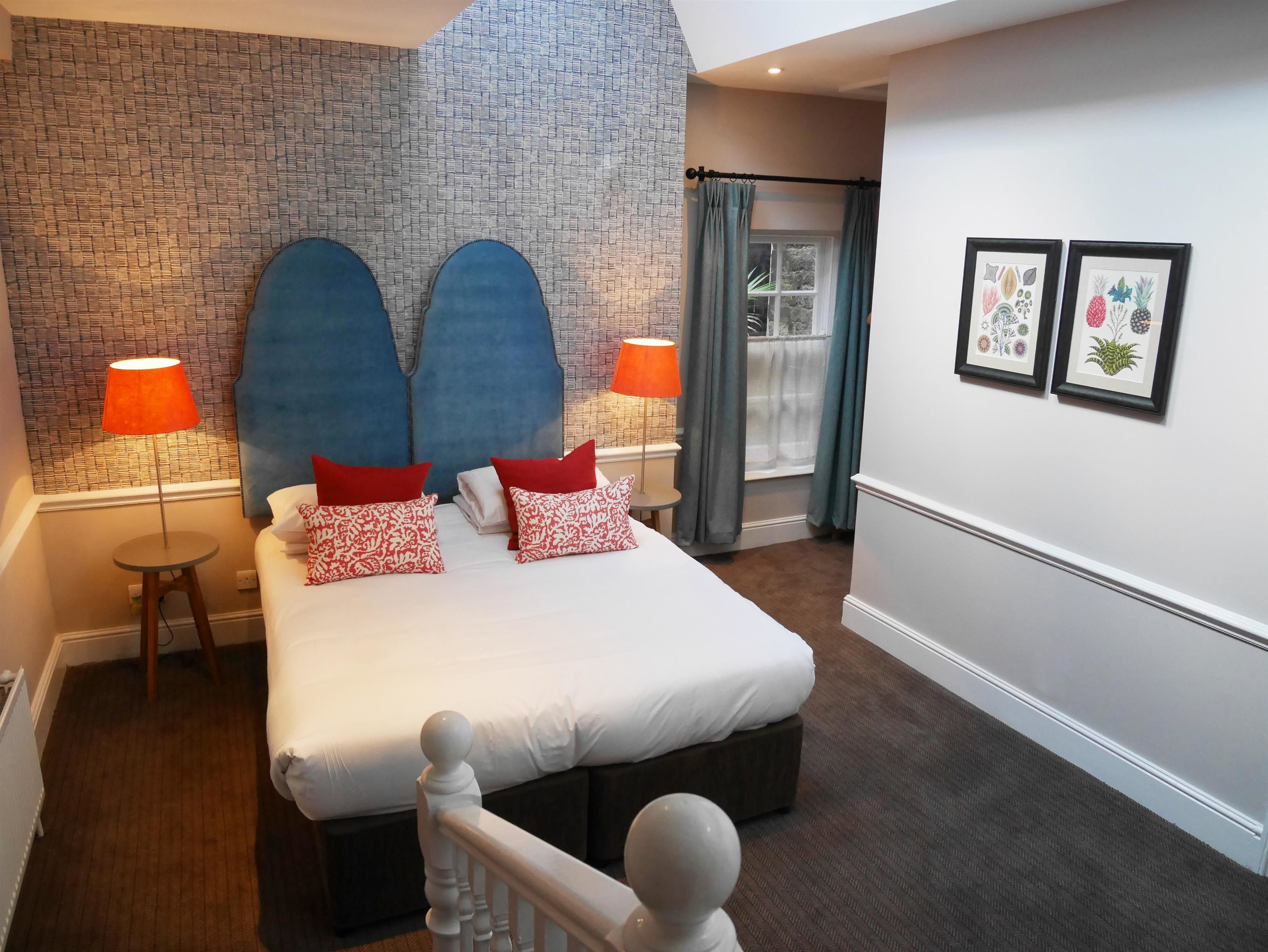 A hotel room with a brick-patterned headboard, a large bed with red and white pillows, two bedside lamps, a small round table, and a window with curtains.