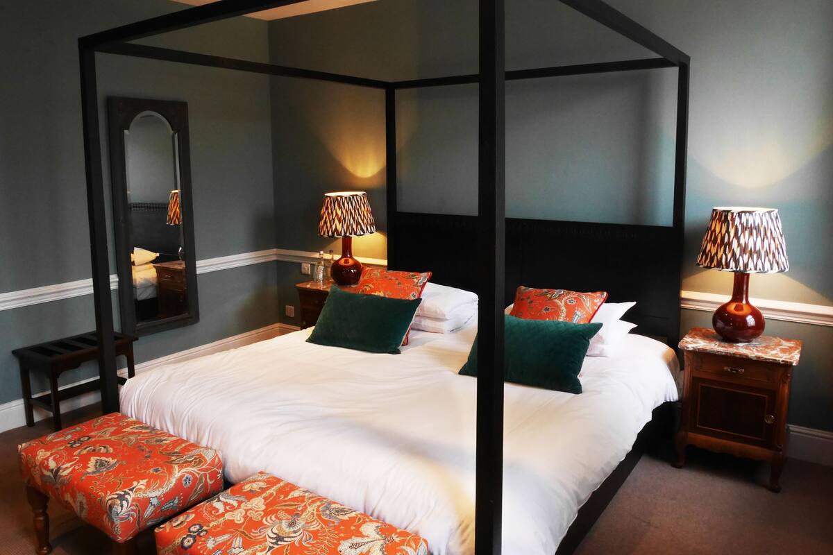A four-poster bed with a canopy, two bedside tables with lamps, a mirror, and a bench with a patterned cushion.