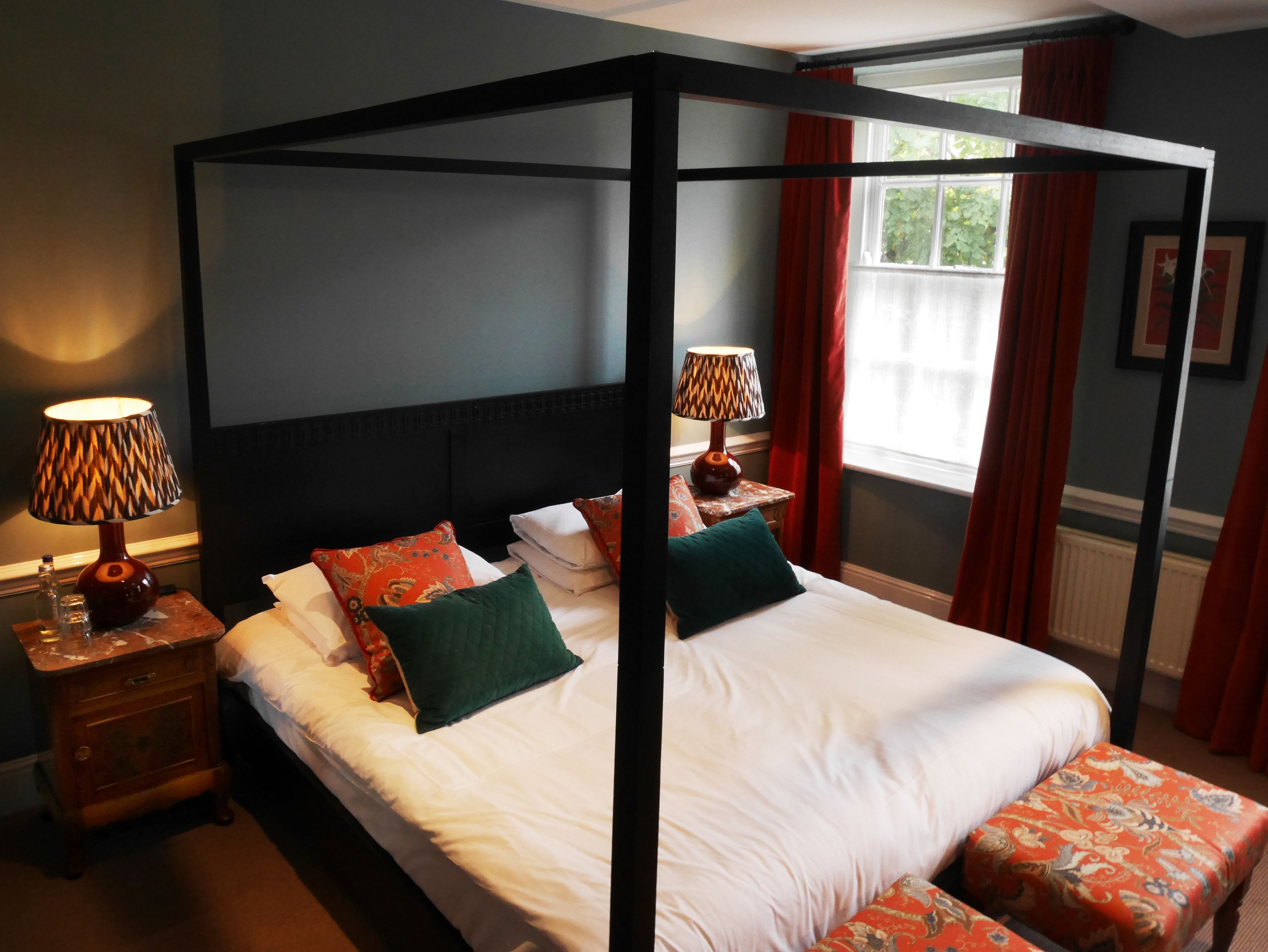 A four-poster bed with white linens and green and red pillows, a nightstand with a lamp, and a window with red curtains.