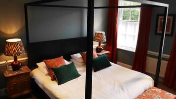 Een hemelbed met witte lakens en groene en rode kussens, een nachtkastje met een lamp, en een raam met rode gordijnen.
