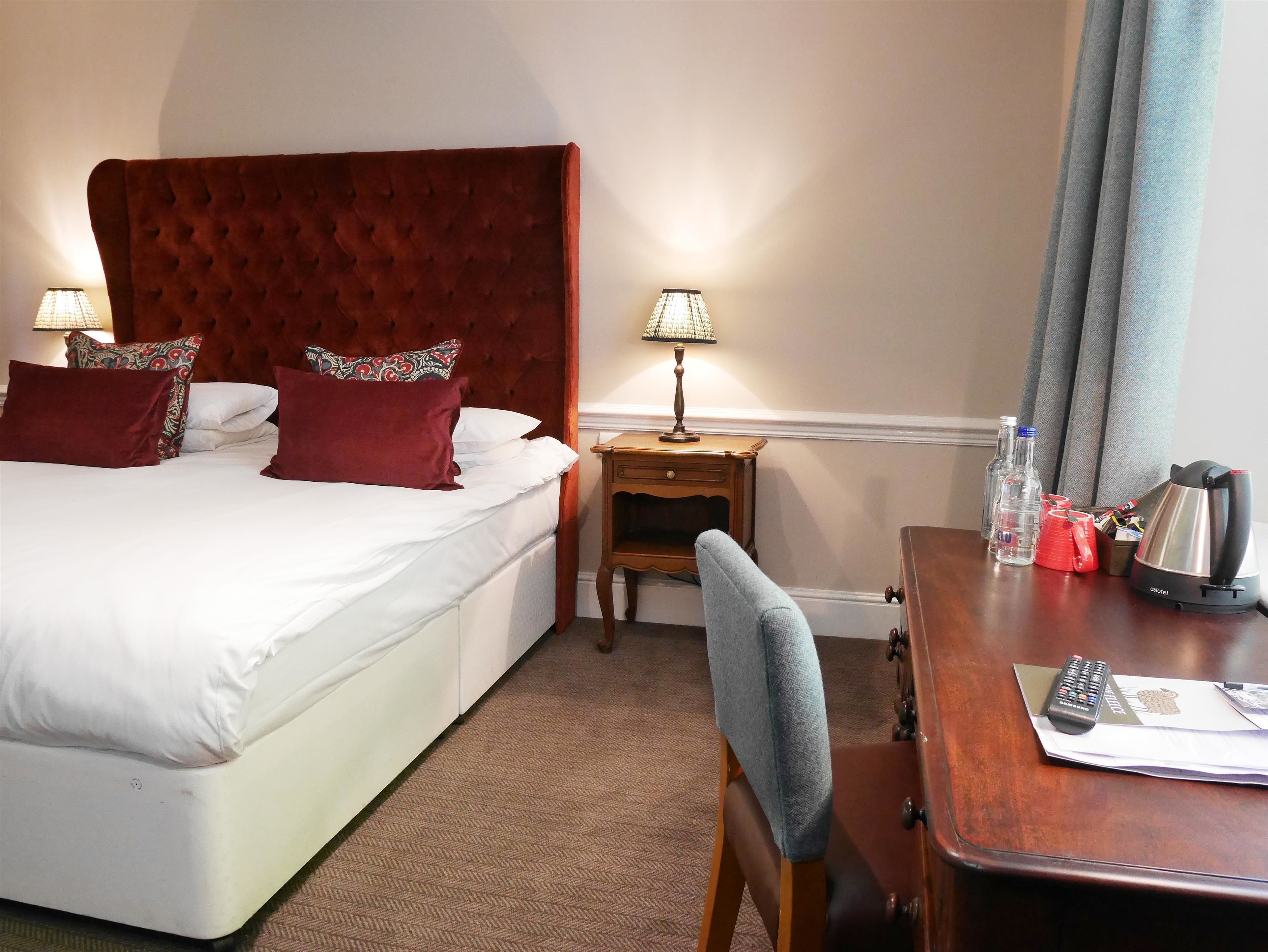 A hotel room with a large bed, two bedside lamps, a wooden nightstand, a desk with a chair, and a kettle.