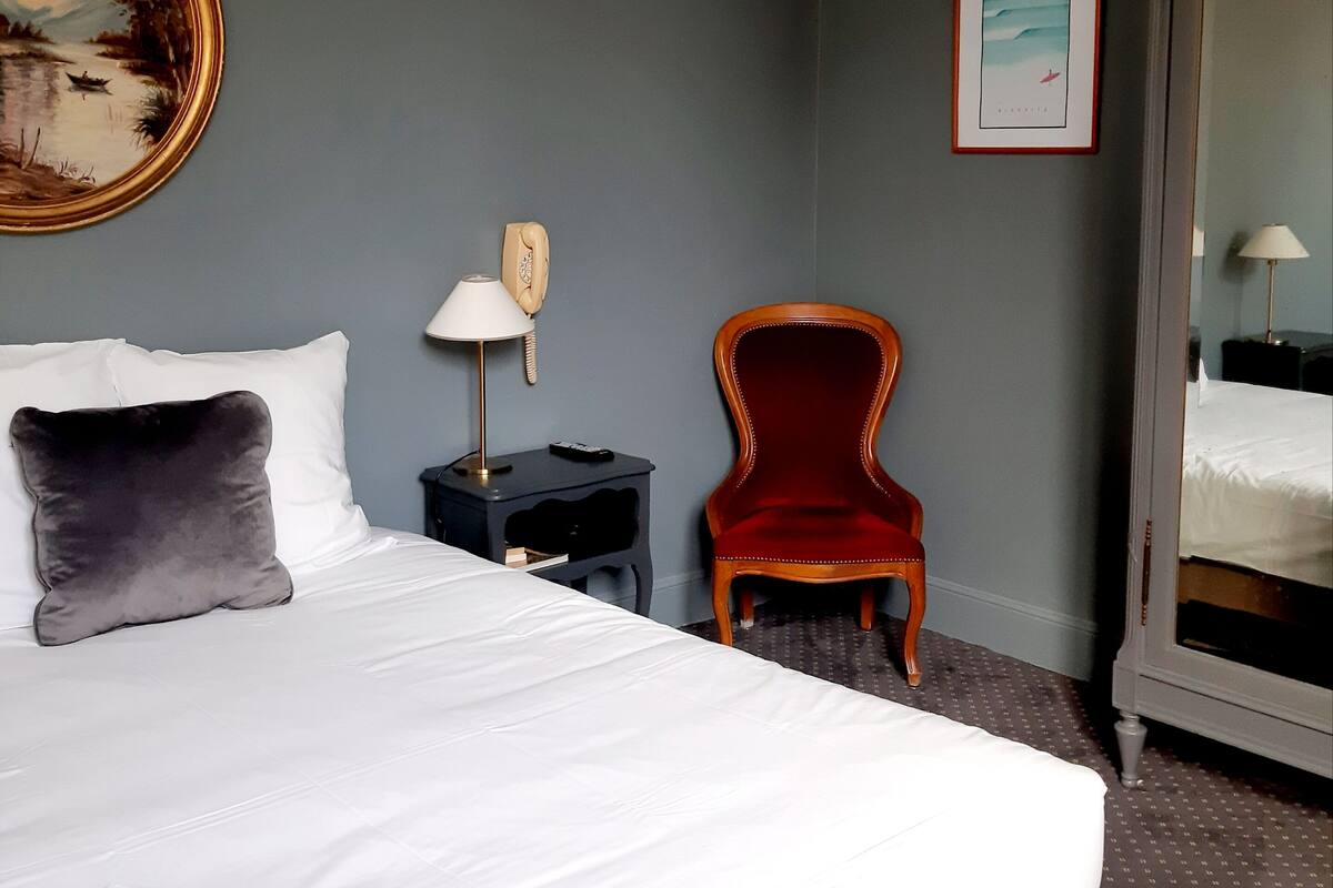 Una habitación de hotel con una cama grande, una mesita de noche con una lámpara, una silla roja y un espejo amplio.