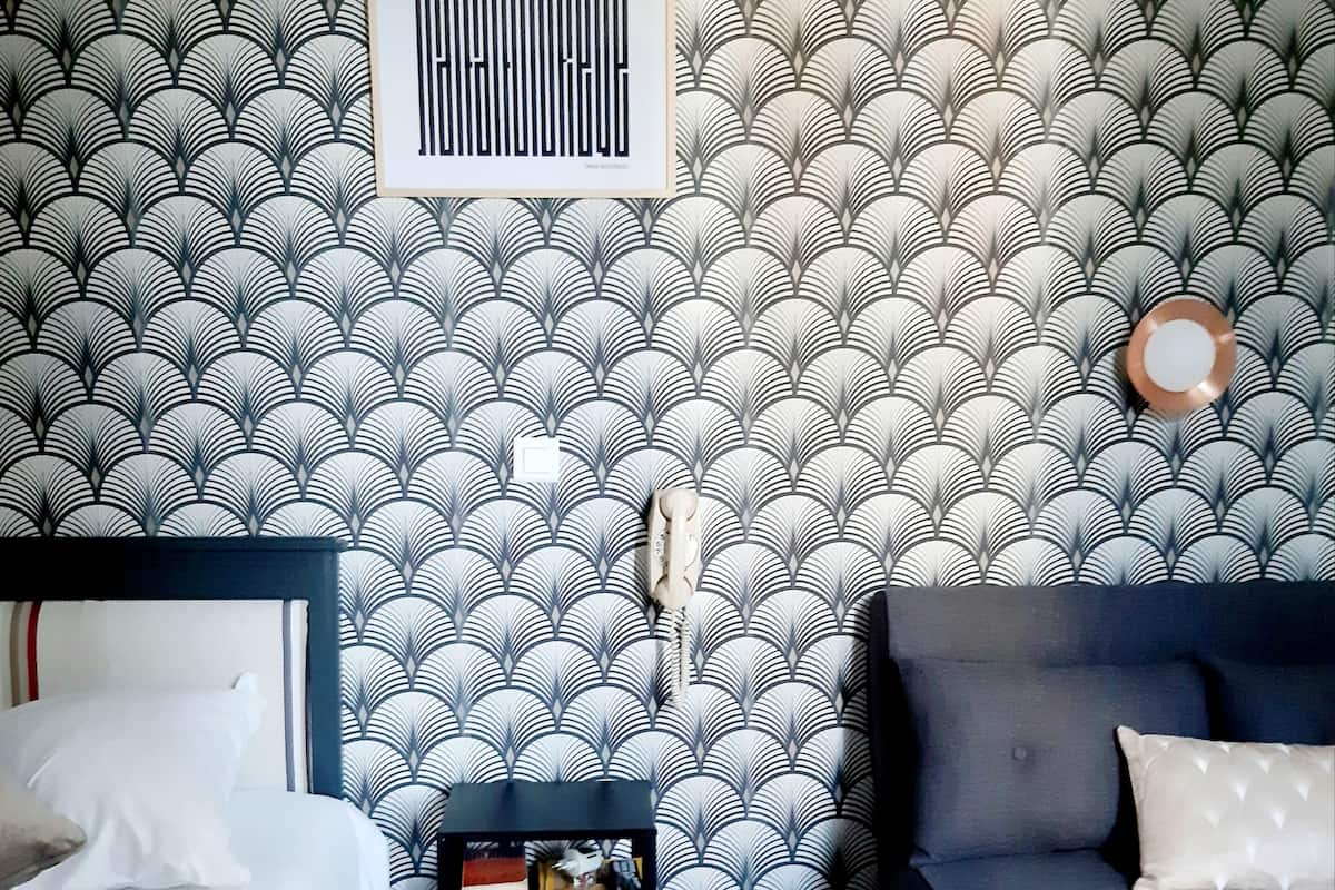 Habitación de hotel con papel tapiz estampado, una cama, una mesita de noche con libros y un sofá gris.