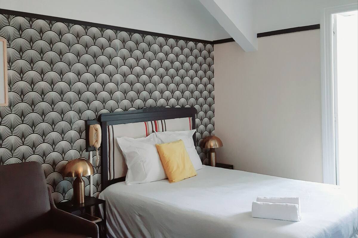 Un dormitorio moderno con papel tapiz estampado, una cama con ropa blanca, un sillón marrón y una lámpara de techo original.