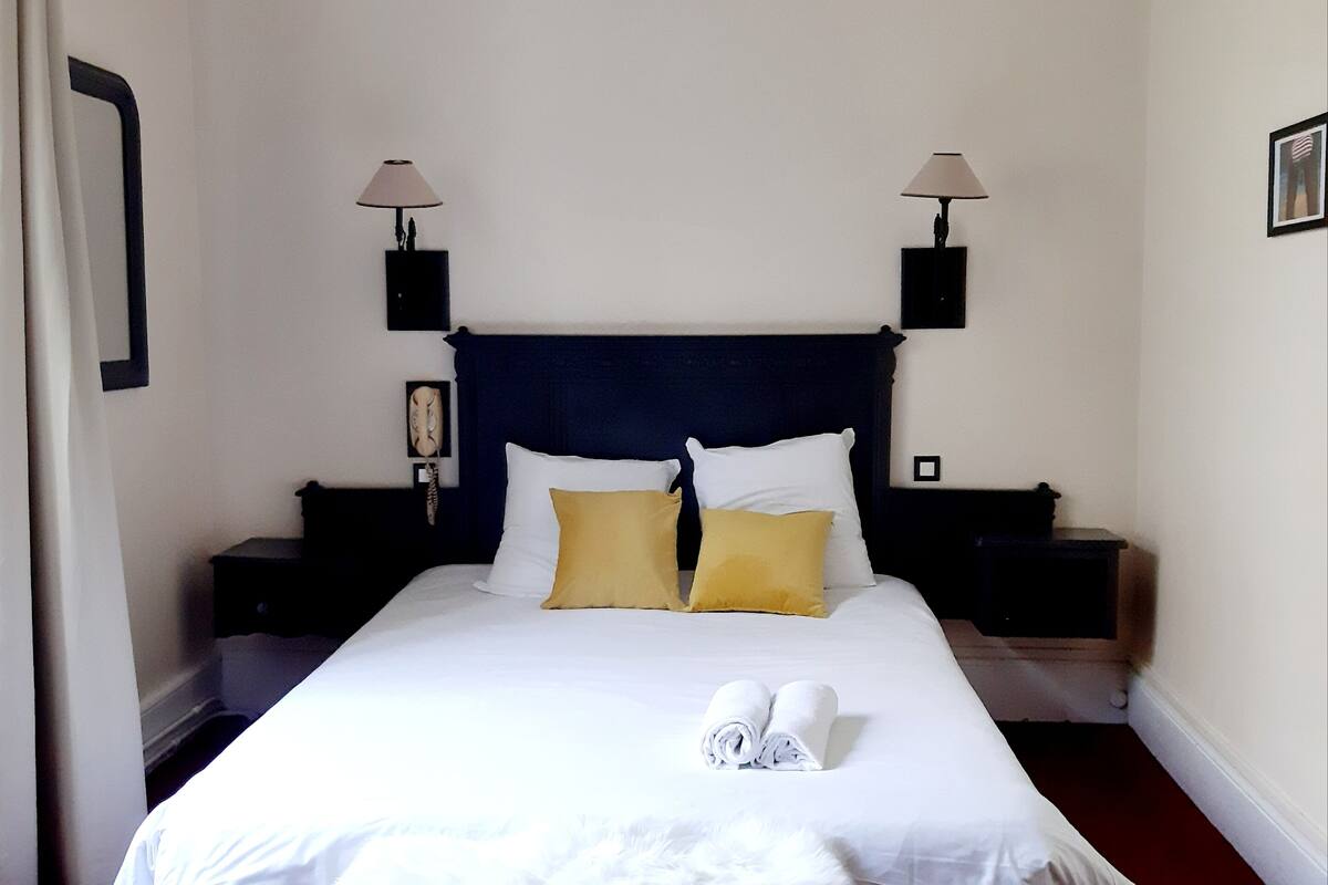 Una cama bien hecha con ropa blanca y dos almohadas, un suave plaid blanco y un par de toallas sobre la cama.