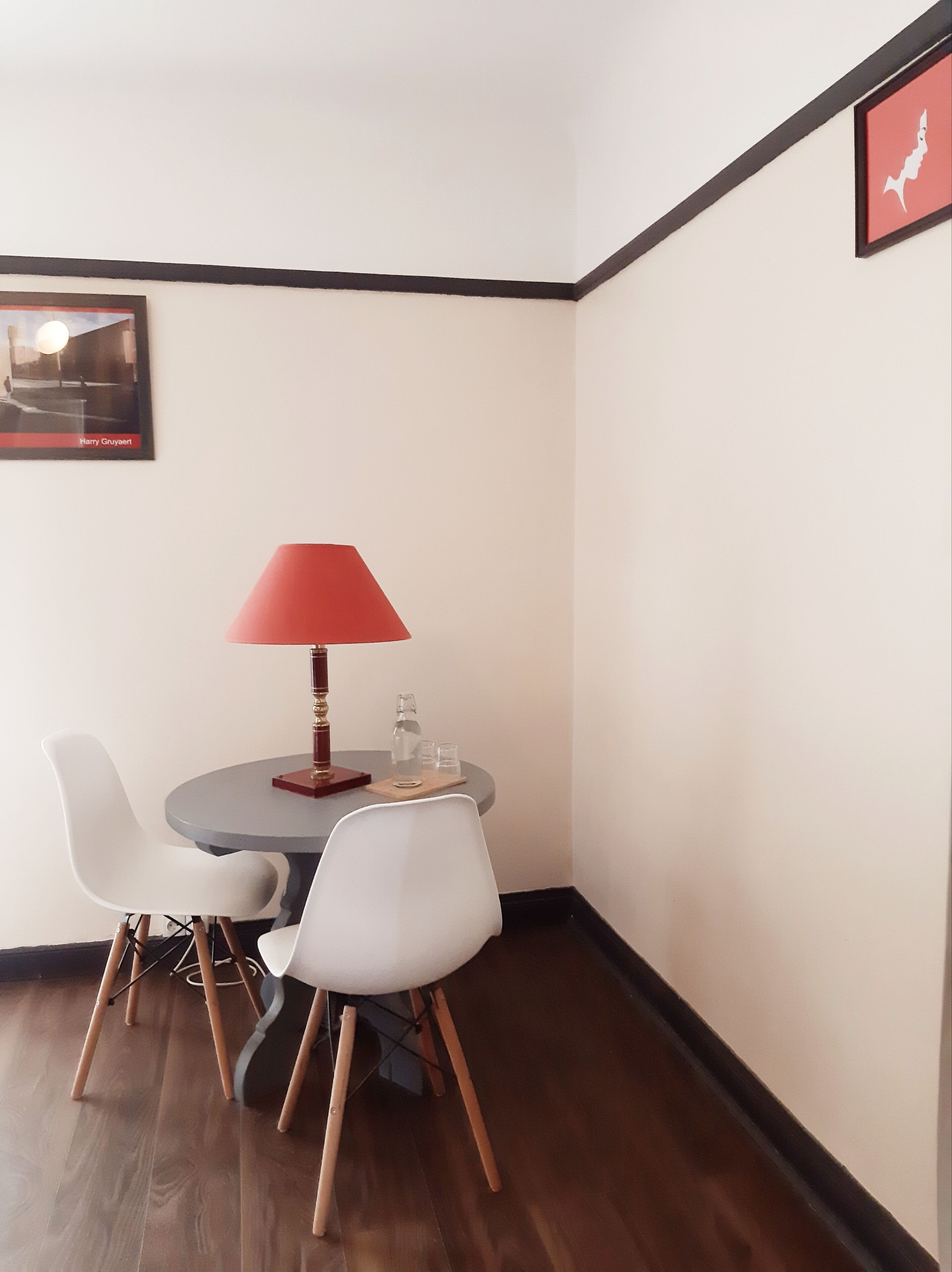 Un pequeño comedor con una mesa redonda, dos sillas blancas, una lámpara de mesa roja y un cuadro en la pared.
