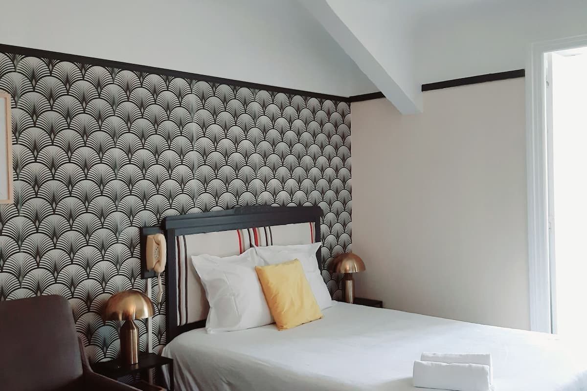 Una habitación de hotel moderna con papel tapiz estampado, una cama con ropa blanca, un sillón marrón, un reposapiés rojo y una lámpara en el techo.
