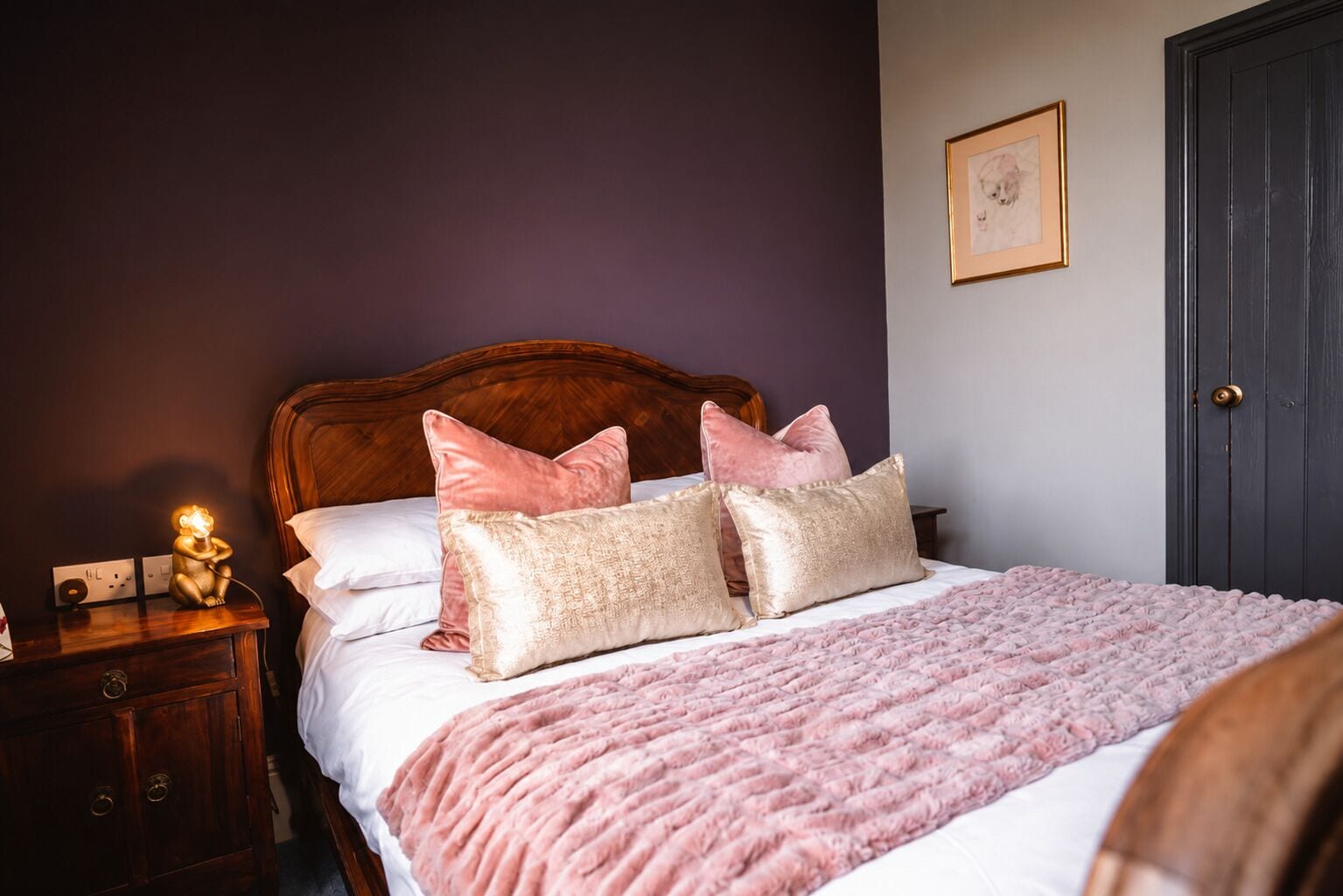 Un dormitorio con cabecera de madera, una cama con almohadas rosa y beige, una manta tejida en rosa y una lámpara de noche, y una puerta.