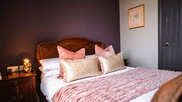Un dormitorio con cabecera de madera, una cama con almohadas rosa y beige, una manta tejida en rosa y una lámpara de noche, y una puerta.