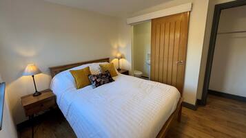 A neatly made bed with white bedding and two pillows, a wooden nightstand with a lamp, and a wooden wardrobe with a door.