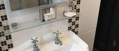 Un baño con un lavabo blanco, dos grifos, un estante de vidrio, un perchero y un espejo.