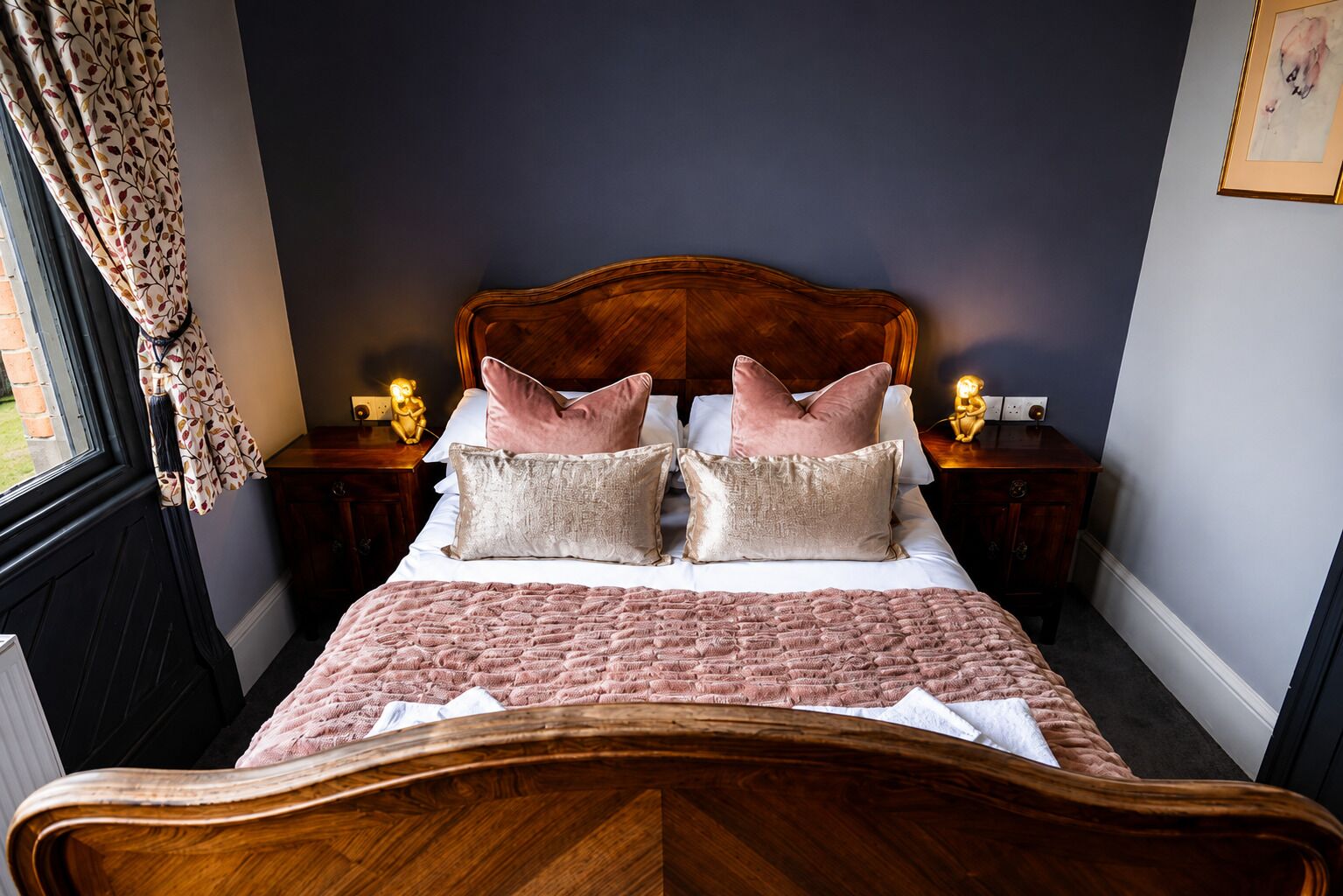 A bedroom with a wooden headboard, a bed with pink bedding, two bedside tables with lamps, and a window with curtains.