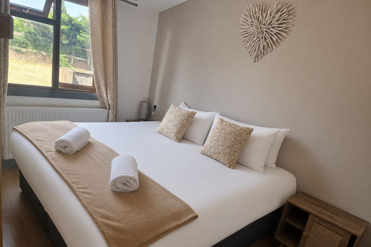 A neatly made bed with a beige blanket and rolled towels, a wooden nightstand, and a decorative wall piece.