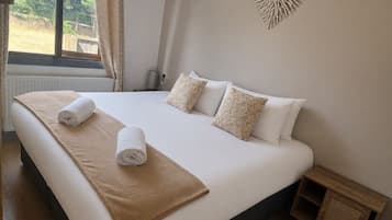 Un letto rifatto con cura, una coperta beige e asciugamani arrotolati, un comodino in legno e un elemento decorativo a parete.