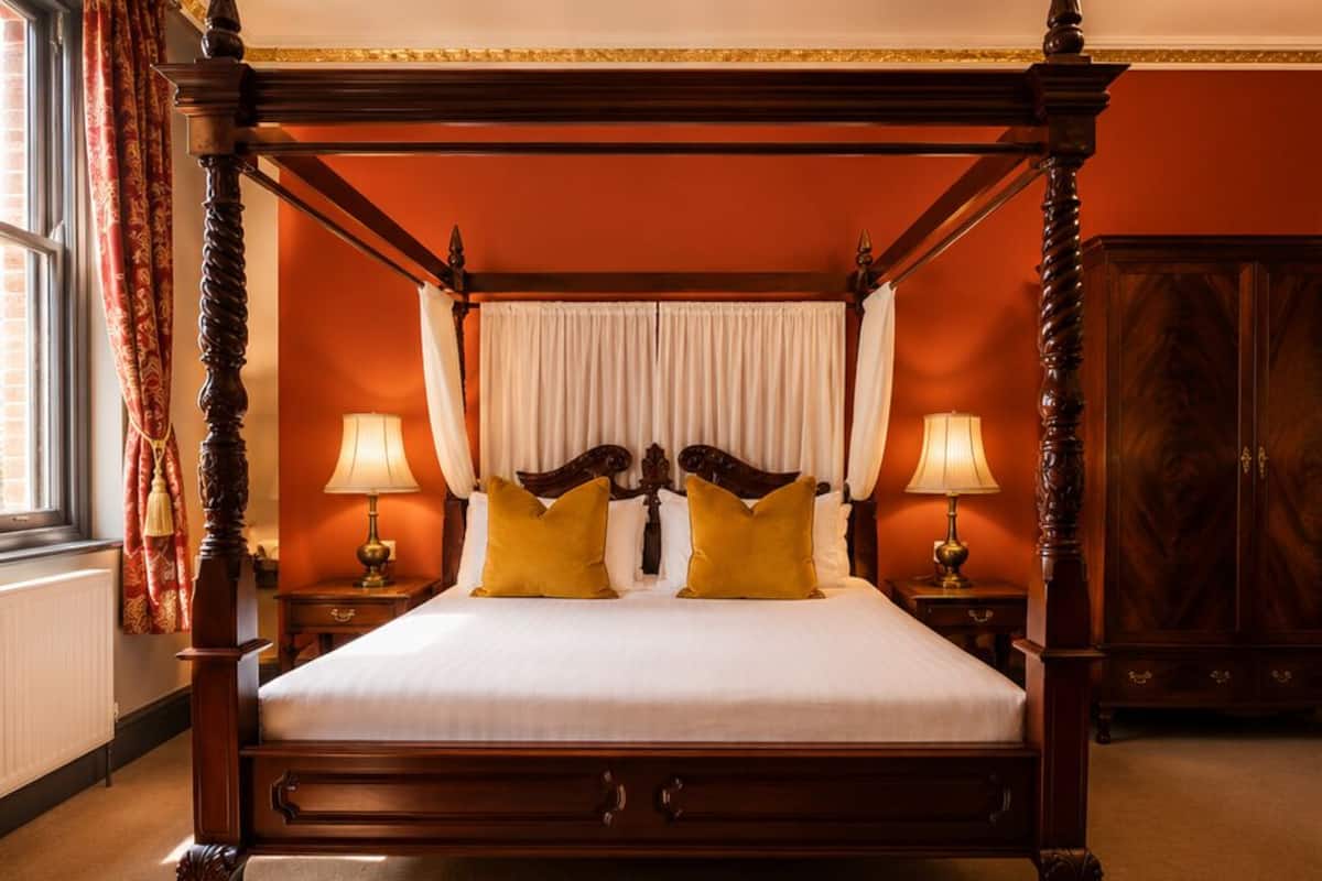 A four-poster bed with white linens and yellow pillows, two bedside lamps, and a wooden wardrobe in a room with orange walls.