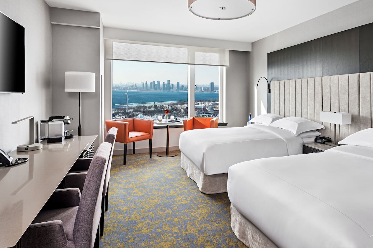 A hotel room with two beds, a desk, a chair, and a view of a city skyline.