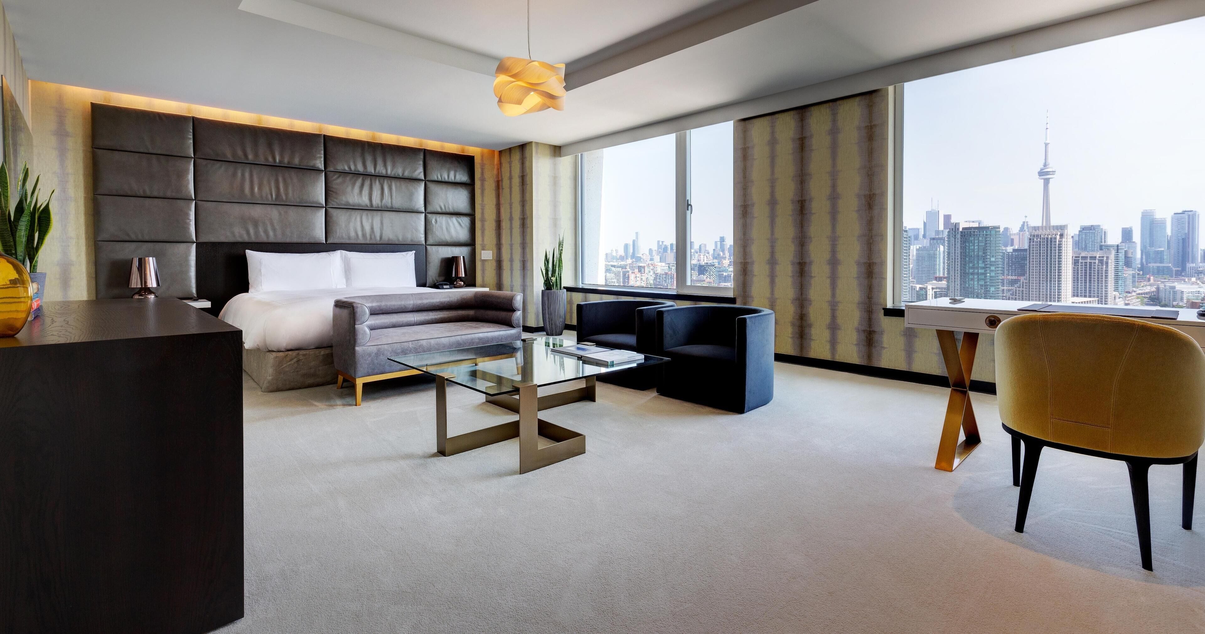 Ein modernes Hotelzimmer mit einem großen Bett, einer Couch, einem Glastisch, einem Schreibtisch und einem Stuhl. Durch große Fenster ist die Stadt zu sehen.