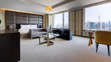 Ein modernes Hotelzimmer mit einem großen Bett, einer Couch, einem Glastisch, einem Schreibtisch und einem Stuhl. Durch große Fenster ist die Stadt zu sehen.
