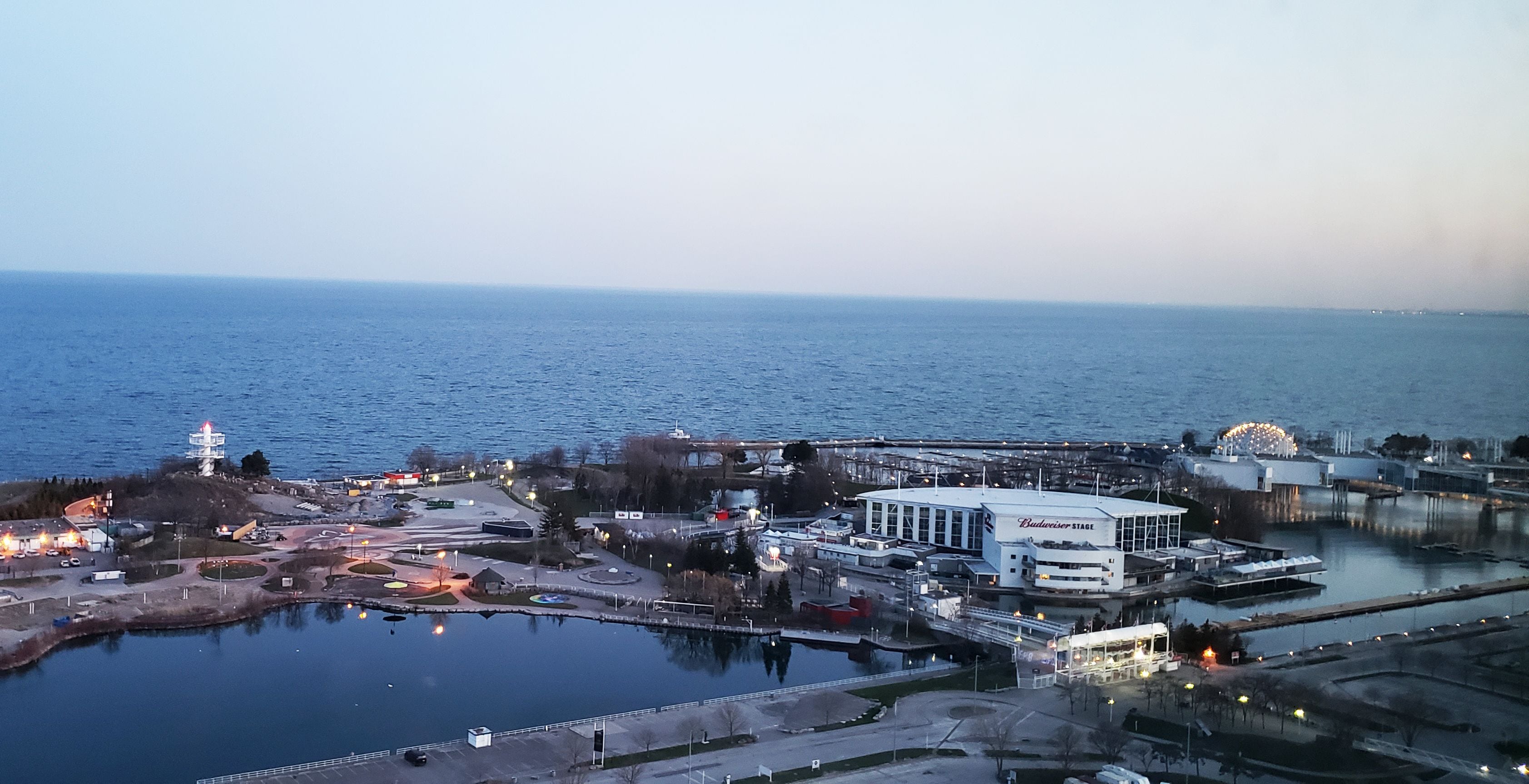 An aerial view of a waterfront area with a large white building labeled "Budweiser Stage," a Ferris wheel, and a marina.
