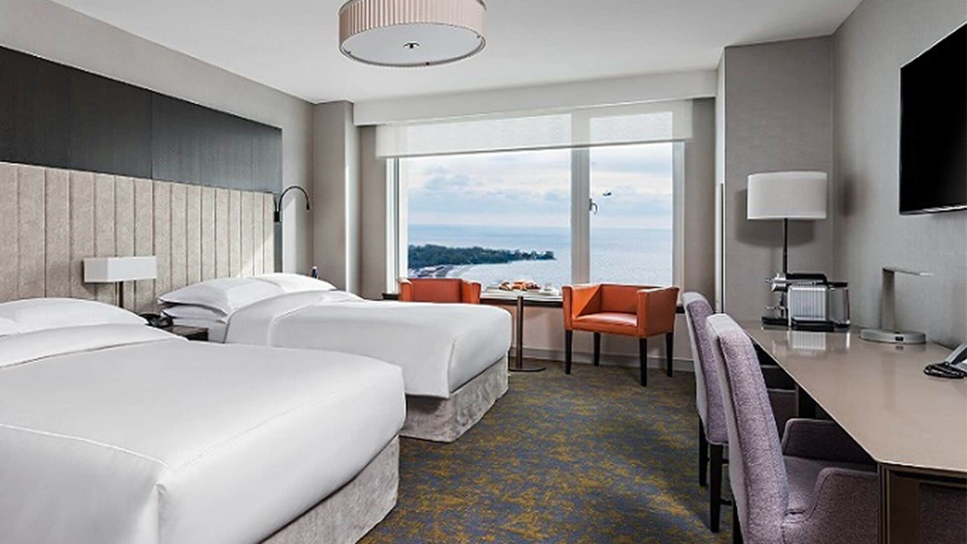 A hotel room with two beds, a desk, a chair, a TV, and a large window with a view of the sea.