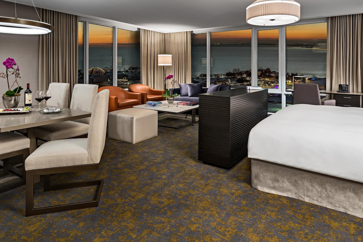 A hotel room with a large window offering a view of the sea, a dining area with a table set for two, and a seating area with a sofa and armchairs.